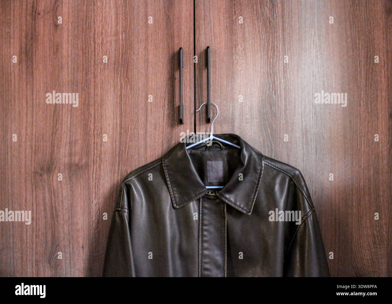 Dark brown retro style leather jacket is hanging on the handle of the wardrobe. Stock Photo