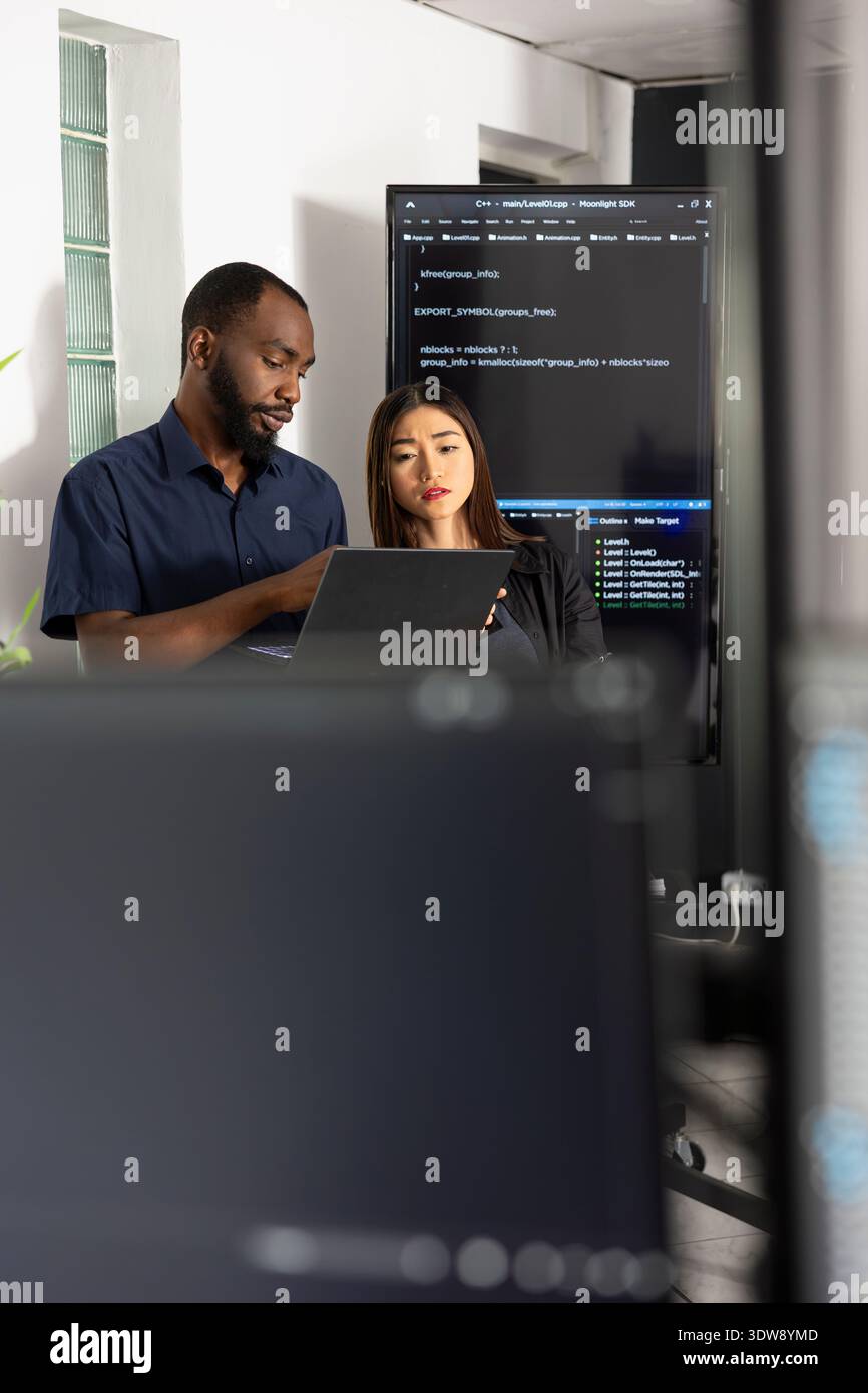 Male and female developers collaborate in tech office, reviewing software data on laptop. Diverse coders analyze HTML scripts together, improving website functionality through teamwork. Stock Photo