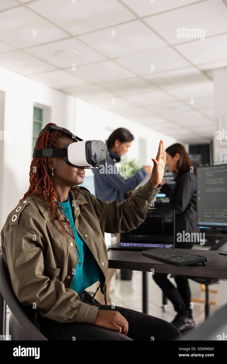 Black female programmer wearing VR glasses interacts with floating 3D interface while working indoors. Futuristic software developer explores augmented reality technology for advanced app development. Stock Photo