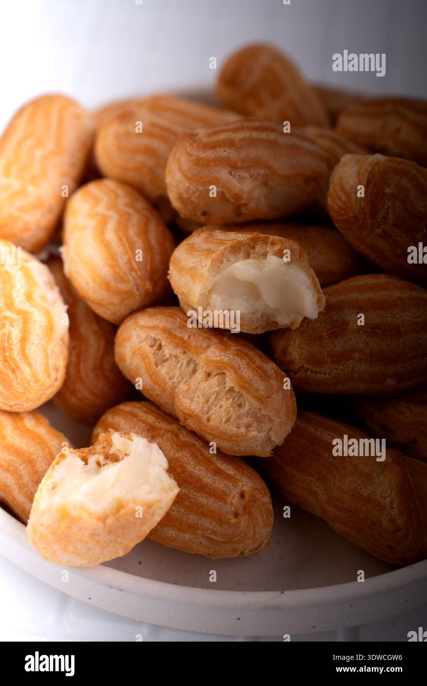 close-up of golden mini eclairs piled in a ceramic bowl: cream-filled choux pastries with ridged glossy shells and soft white filling, warm natural li Stock Photo