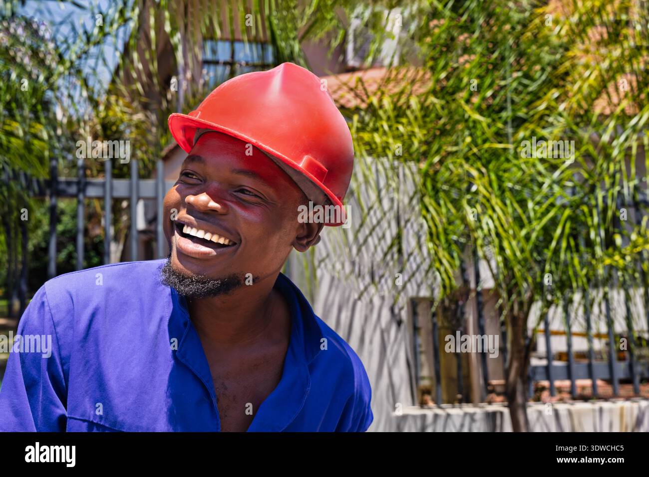 smiling Black man wearing a blue shirt and a red hard hat is captured in a low-angle, hero shot, worker close-up portrait outdoors Stock Photo