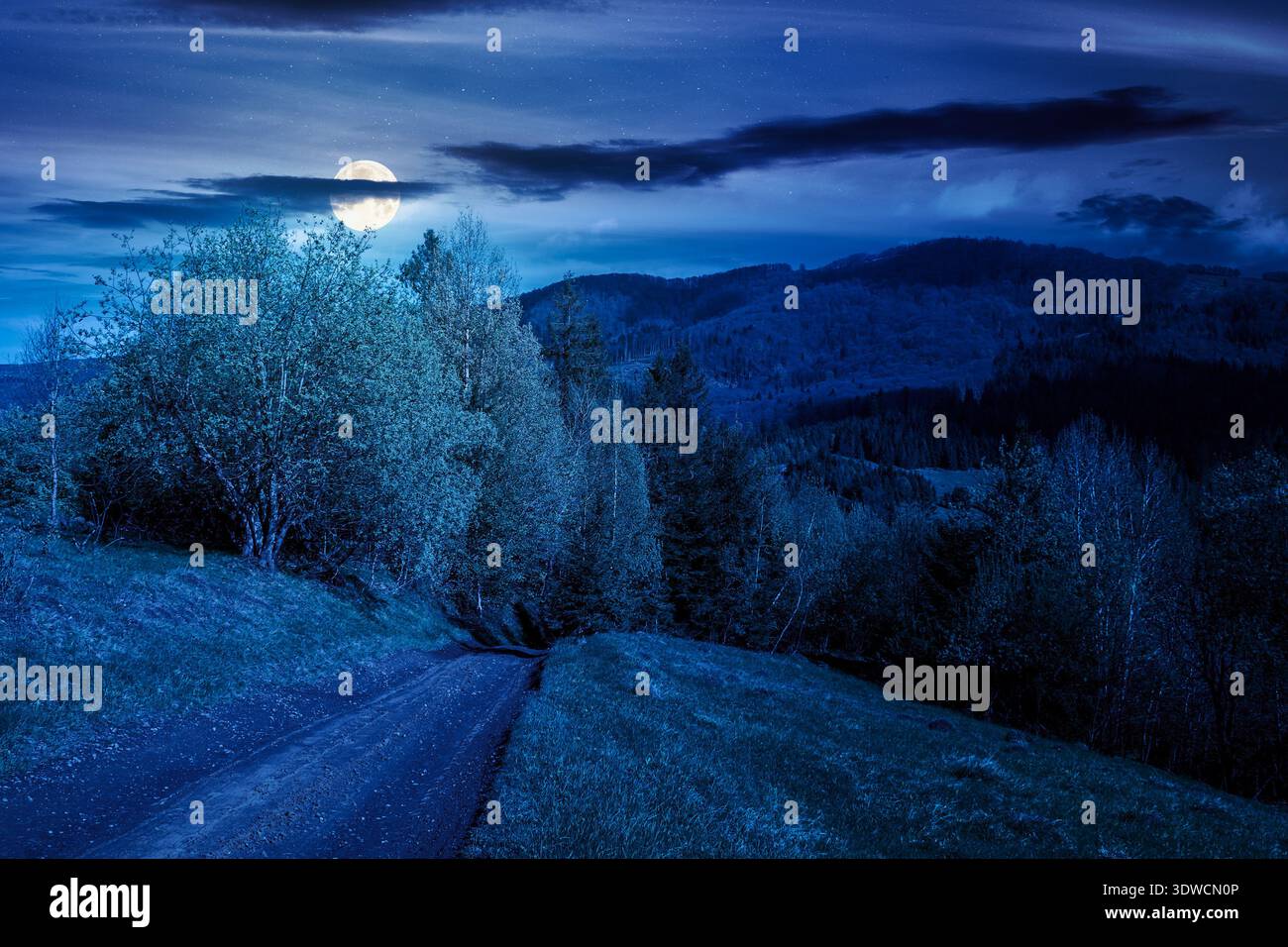 mysterious mountain landscape with dirt road at night. full moon over forest path through grassy hills in spring. rural transportation in greenery sce Stock Photo