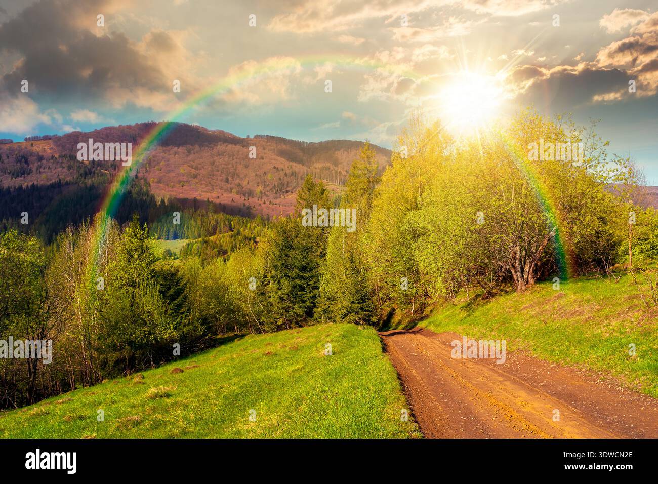 sunset over beautiful mountain landscape with dirt road. forest path through grassy hills in spring. rural transportation in greenery scenery. empty s Stock Photo