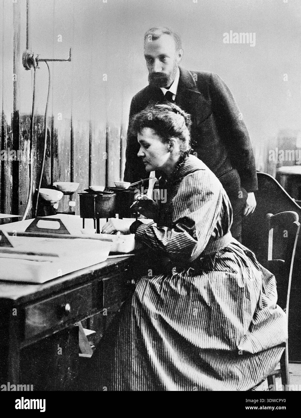 This vintage black and white photograph captures Nobel Prize winners Marie Curie and Pierre Curie working together in their laboratory, likely in the late 1890s or early 1900s. Marie is seated at a desk, engaged in scientific work with laboratory equipment, while Pierre stands nearby, observing or assisting her. Stock Photo