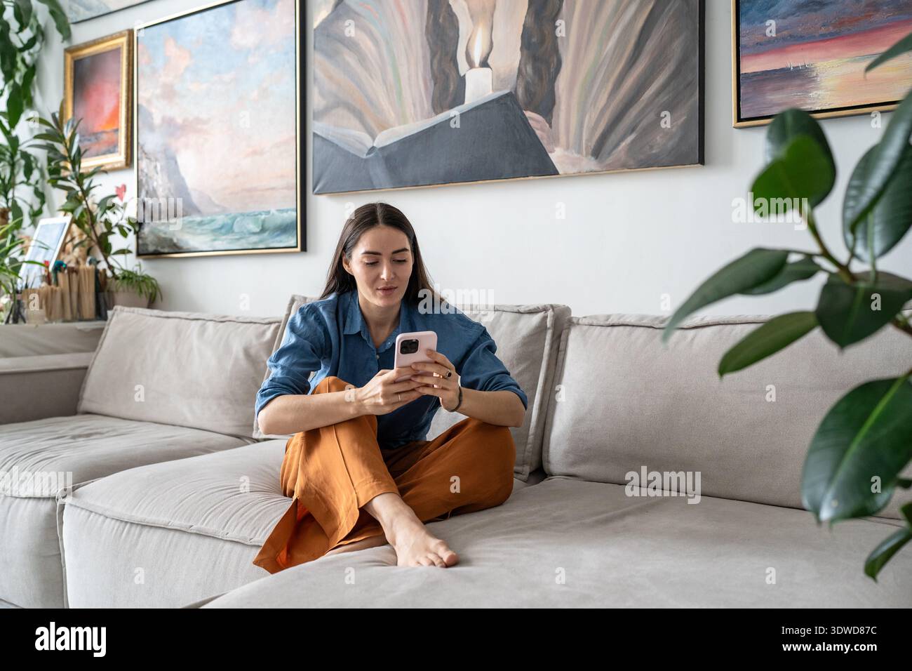 Curious woman artist sitting on couch watching media online on smartphone at cozy home gallery. Stock Photo