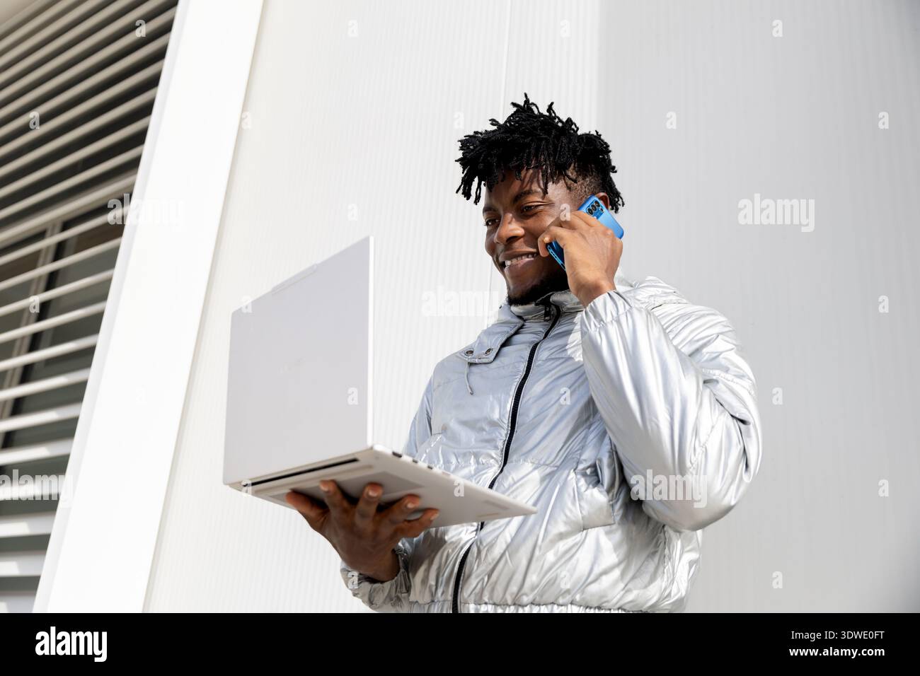A man wearing a shiny silver jacket is standing outside, holding a laptop in one hand while talking on a smartphone. The setting appears modern and fu Stock Photo