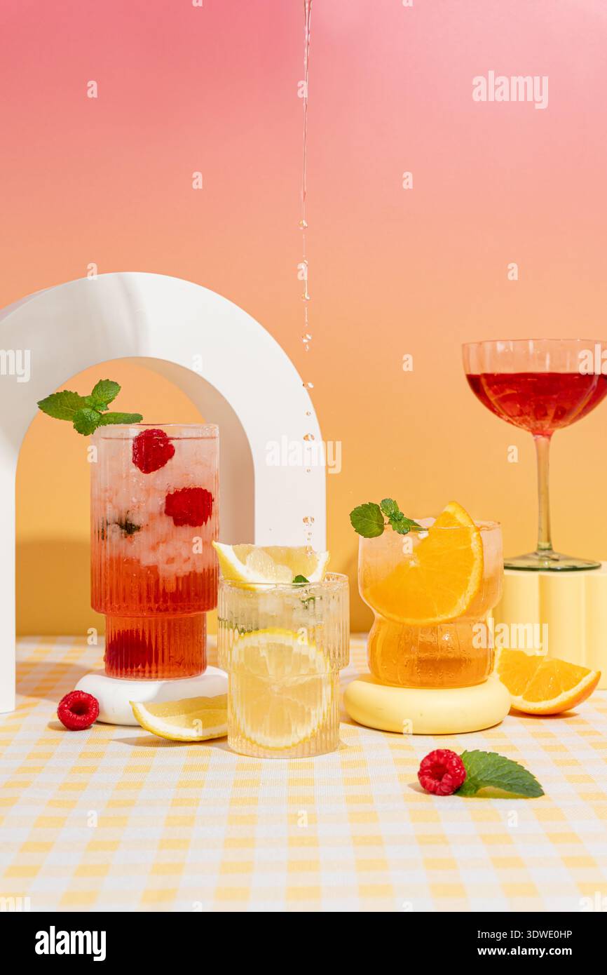 A vibrant display of cocktails featuring raspberries, oranges, and lemons, set against a pink and orange ombre background on a yellow checked tableclo Stock Photo