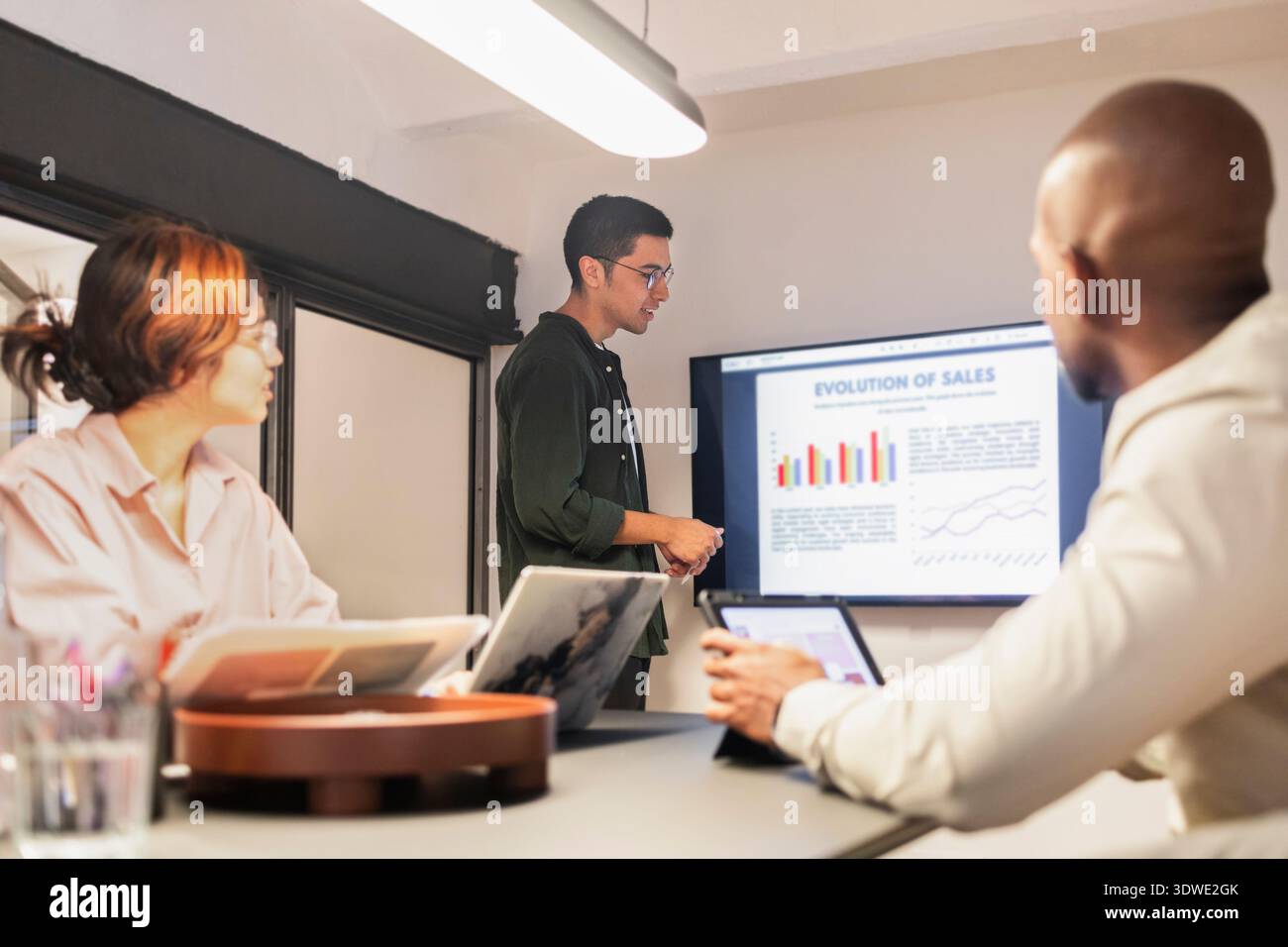 Diverse team engaged in a meeting at a coworking office. A presenter discusses sales graphs on a digital screen while colleagues attentively follow wi Stock Photo