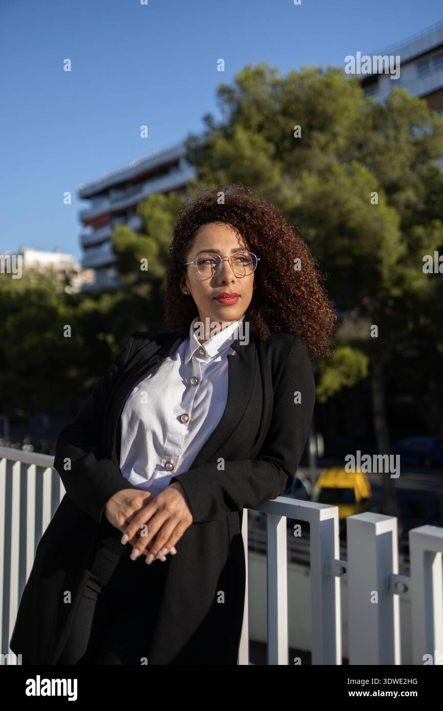 A confident business woman with curly hair stands outside, exuding professionalism. She wears glasses and is dressed in a smart business suit, leaning Stock Photo