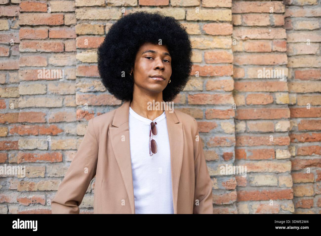 A stylish person stands confidently against a rustic brick wall, wearing a brown blazer over a white shirt, with sunglasses hanging from their shirt c Stock Photo