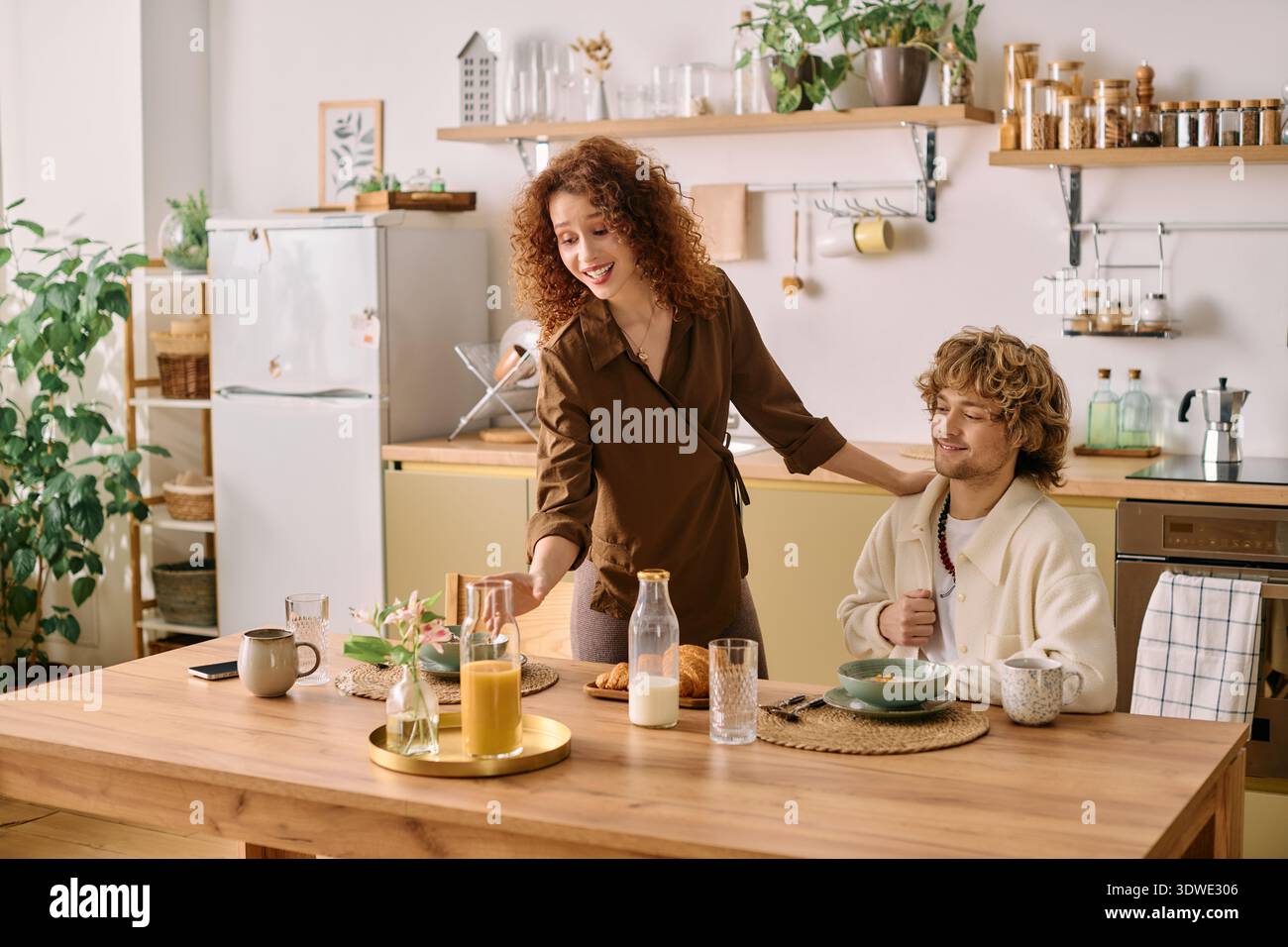 Redhead woman serves breakfast to her blonde partner in their stylish kitchen setting. Stock Photo