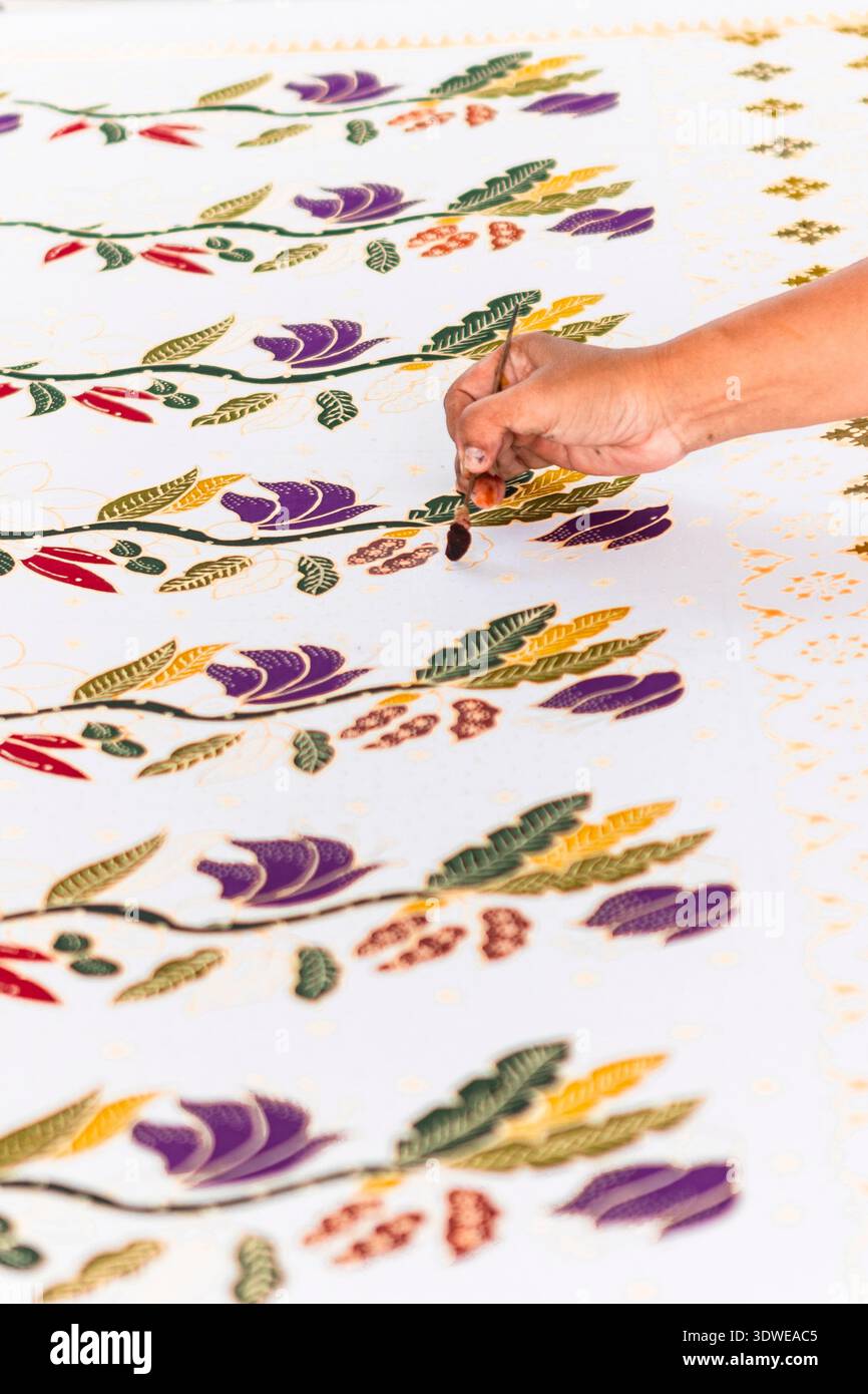 A close-up of a skilled artisan's hand applying traditional Indonesian batik hand-dyeing techniques on fabric. The intricate patterns feature vibrant Stock Photo