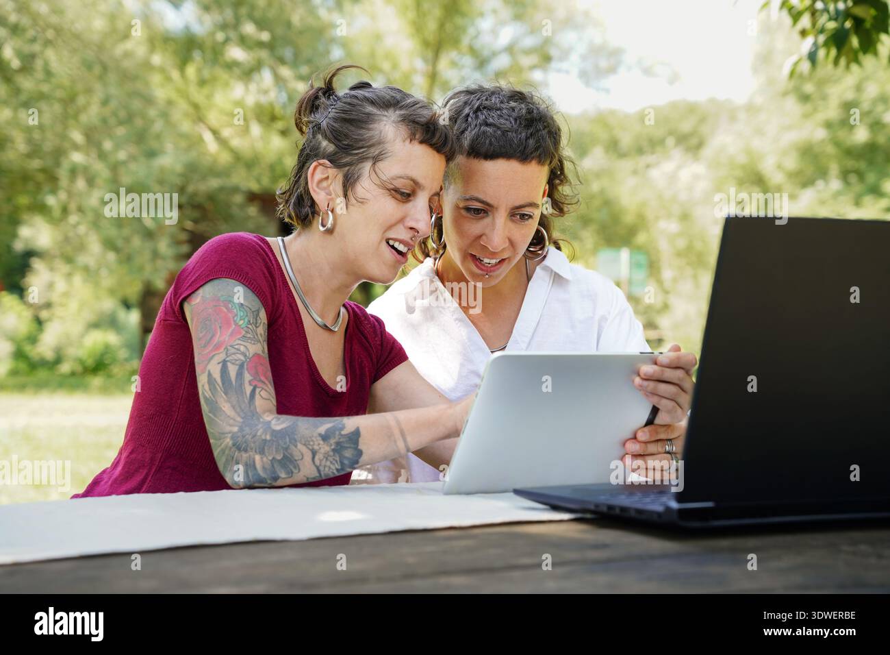 Two sisters enjoying a delightful day outdoors, engaging in digital activities. They are smiling and sharing information using a tablet, surrounded by Stock Photo