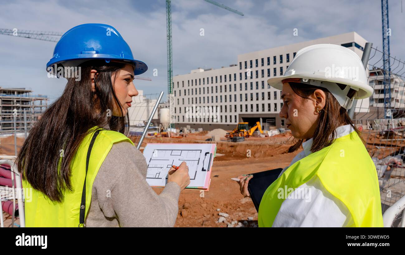 An architect collaborates with a technical engineer on a construction site, examining blueprints and discussing project details. Both wear safety gear Stock Photo