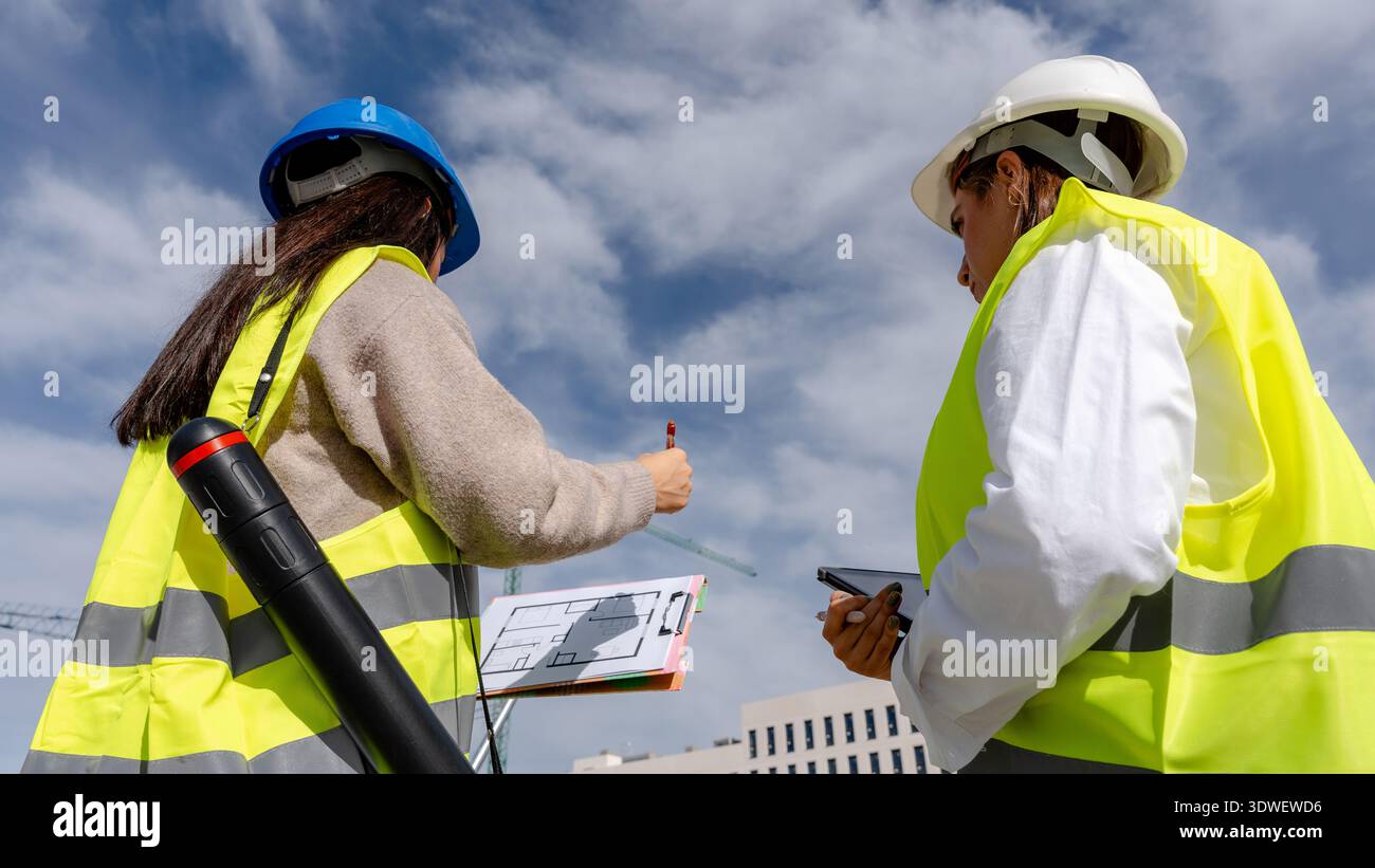 An architect and a technical engineer wearing safety gear discuss blueprints on a construction site. They analyze plans collaboratively, ensuring proj Stock Photo