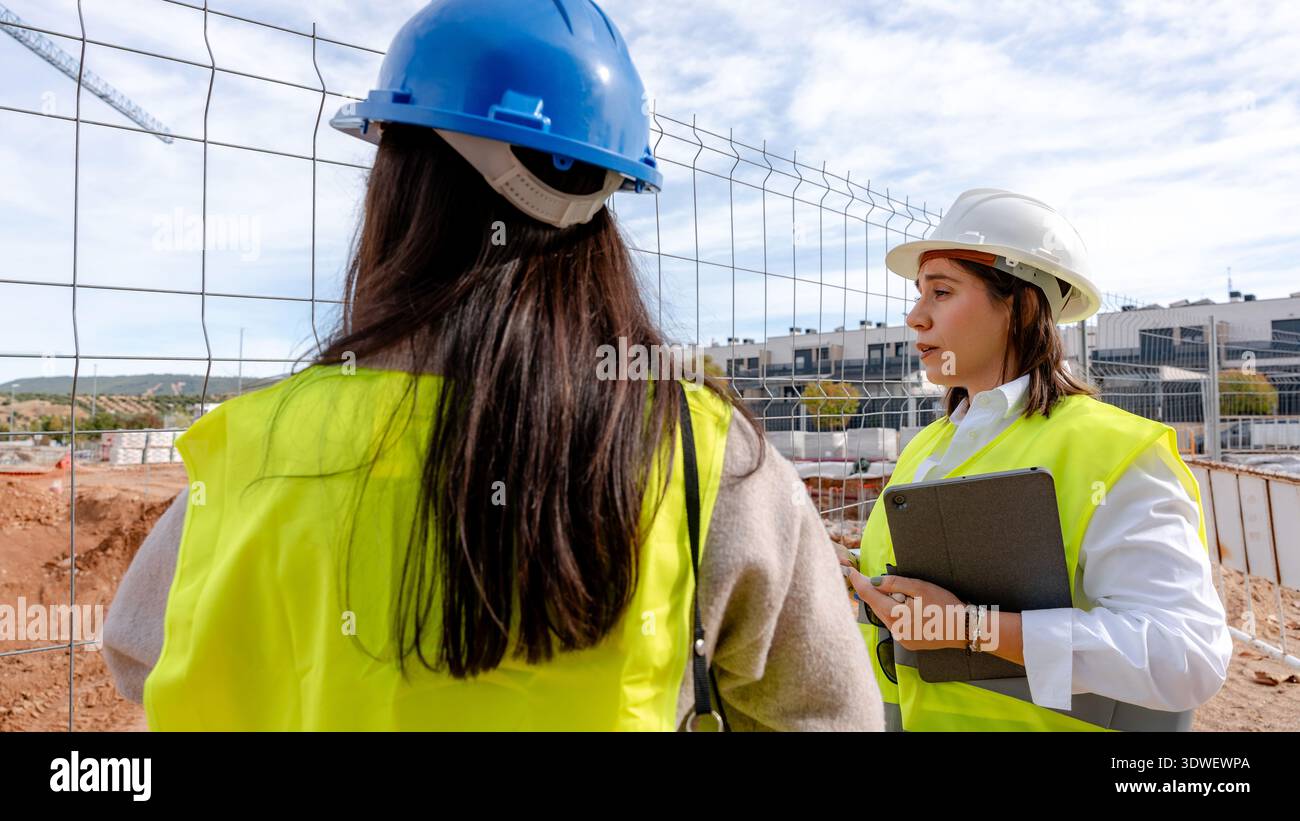 An architect and a technical engineer discuss plans on a construction site. Both are wearing safety helmets and high visibility vests, examining archi Stock Photo