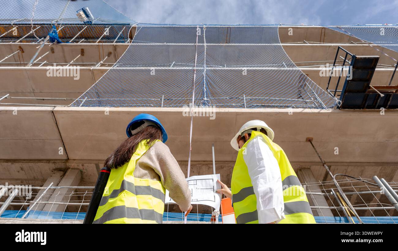 An architect and a technical engineer review blueprints on a construction site, collaborating on building plans. Both are wearing safety gear and obse Stock Photo