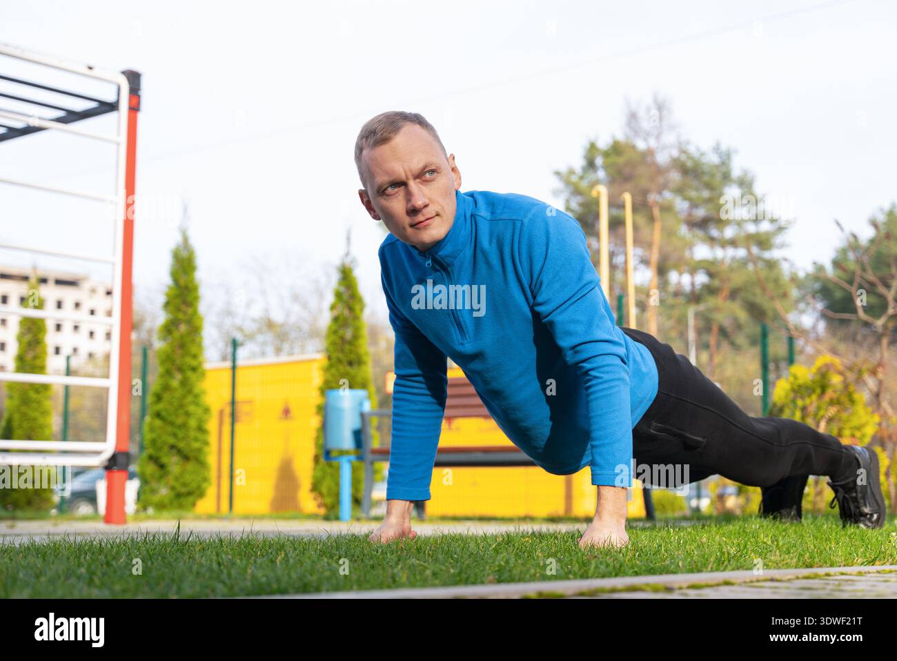 A man in a blue jacket performs outdoor exercises on grass in a park. He maintains good form with a focused expression, surrounded by green trees and Stock Photo