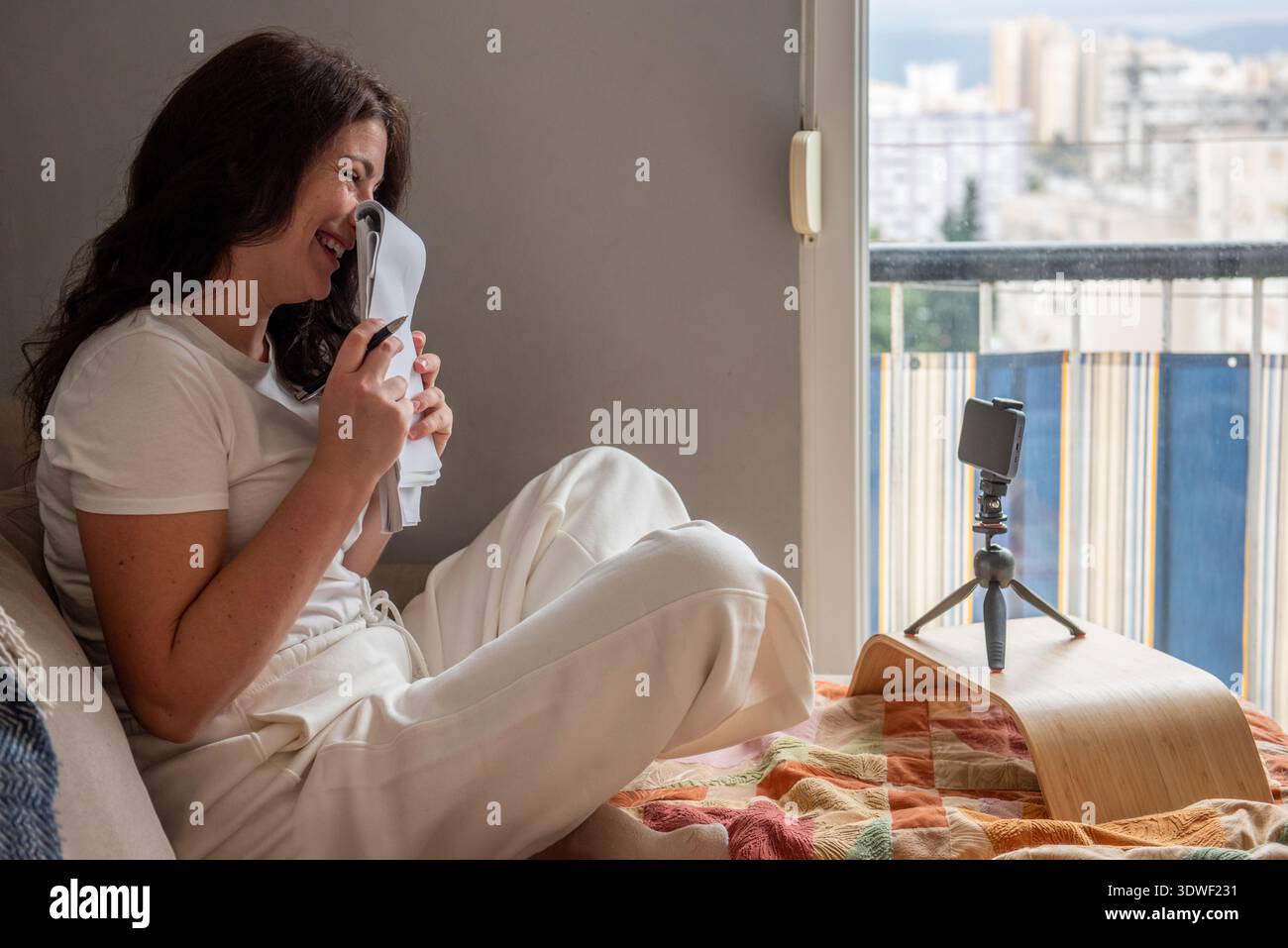 A woman is blogging from home, streaming live using her smartphone on a tripod. She is sitting comfortably, creating engaging content for her audience Stock Photo