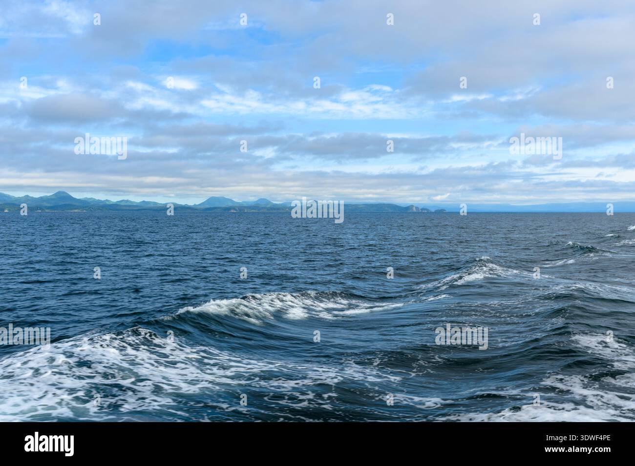 Soft waves create ripples across the ocean, while clouds drift above. Distant mountains rise against the horizon, reflecting a tranquil coastal scene. Stock Photo