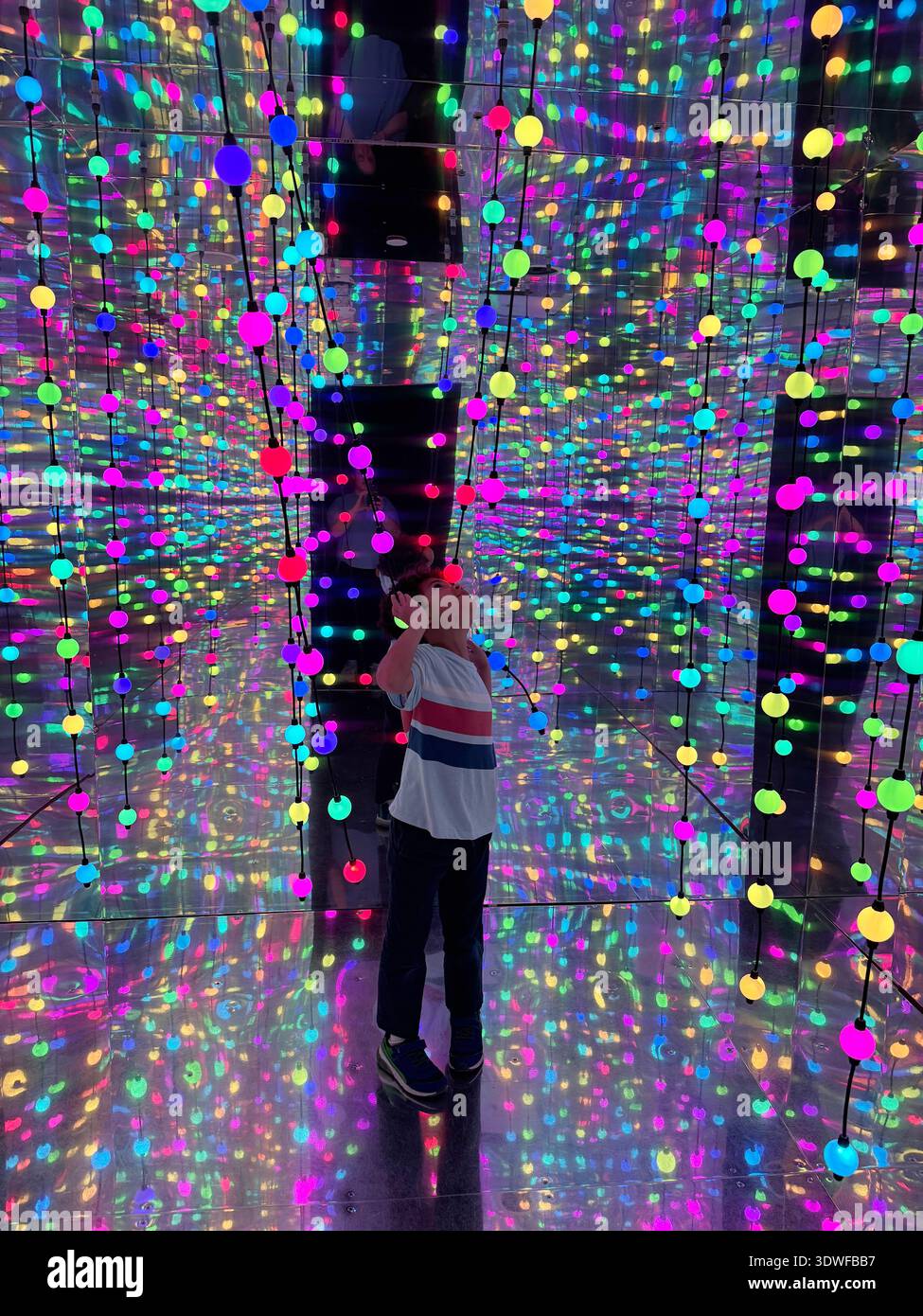 Happy child in a mirrored infinity room filled with hanging multicolored LED lights, creating a vibrant immersive light installation. - Smartphone Captured Stock Image