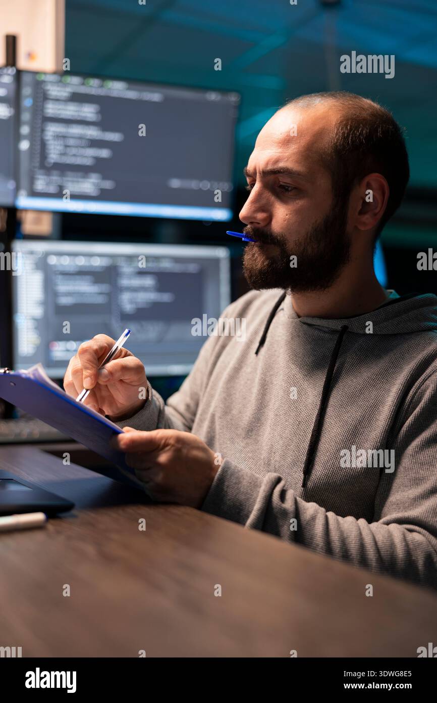 Programmer reviews paperwork with pen cap held between lips, checking coding scripts on laptop. Developer analyzes technical details on clipboard and confirms information against data on device. Stock Photo