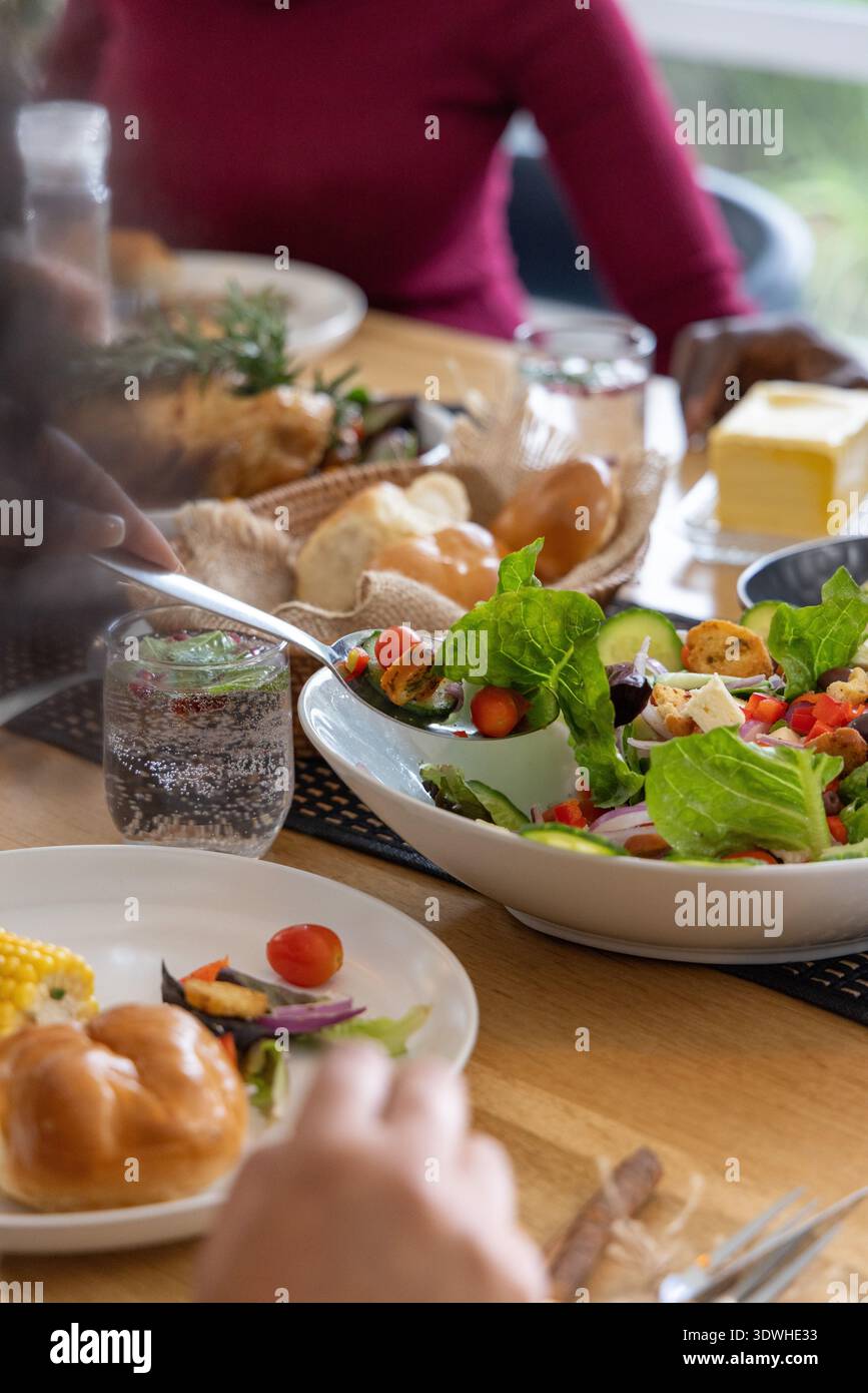 Diverse friends sharing meal at wooden table, reaching for white salad bowl, bread basket and corn Stock Photo