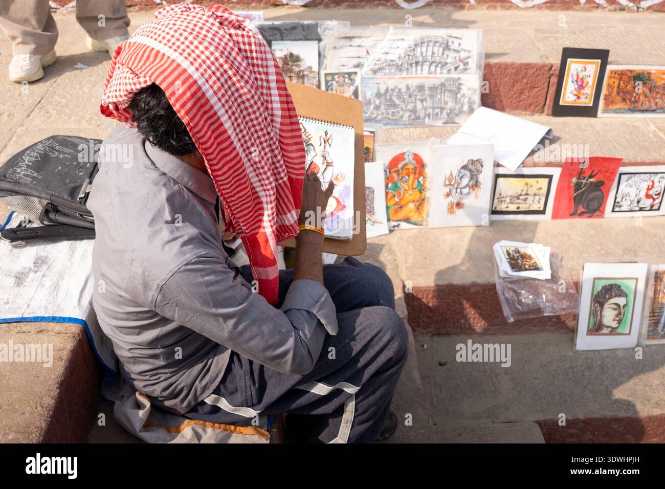 Artist painting Hindu religious artwork on the streets of Varanasi, India, surrounded by various colorful paintings. Stock Photo
