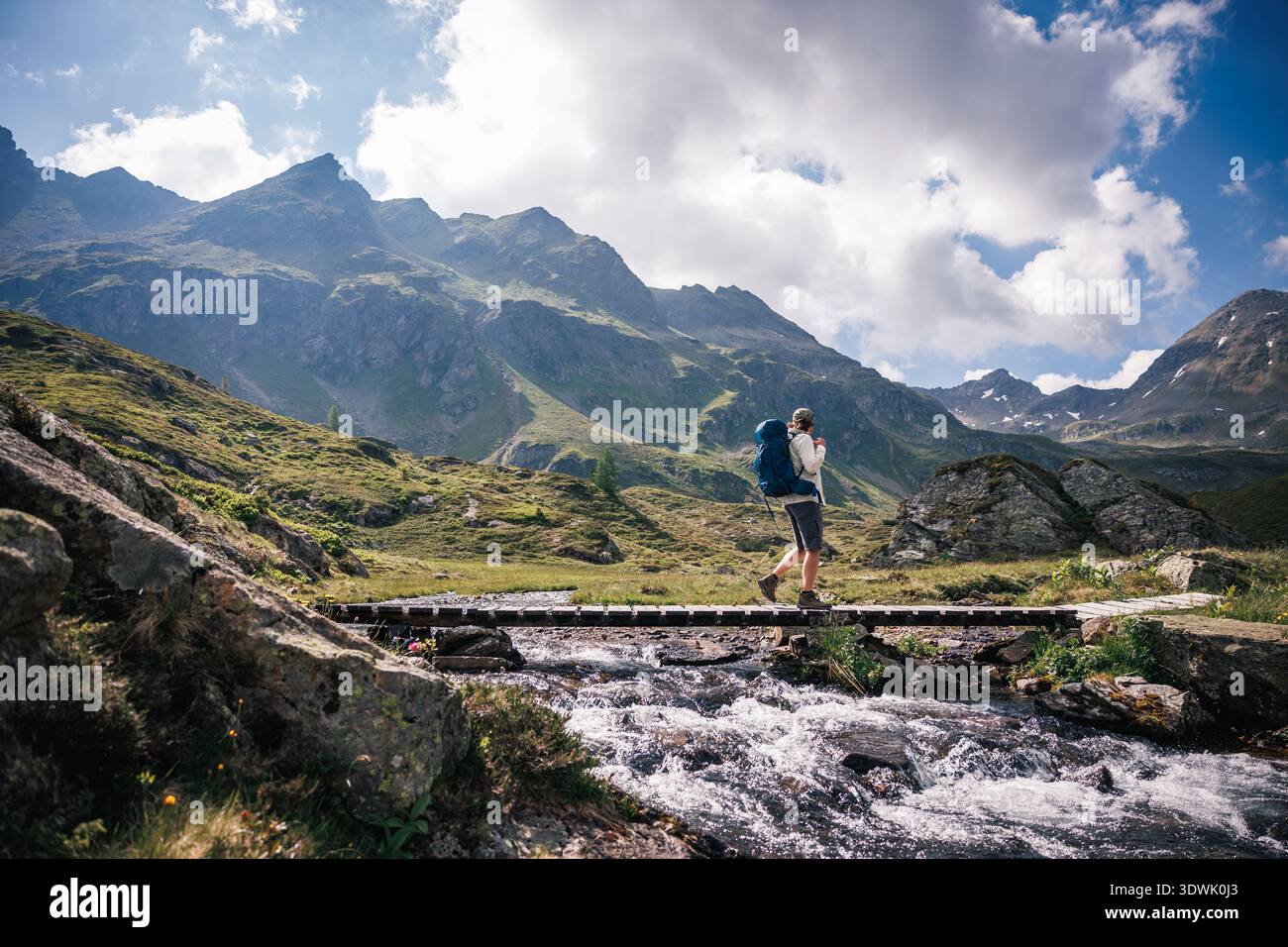 Backpacker crossing wooden bridge over mountain stream on hiking trail in Austrian Alps mountains. Solo hike adventure Stock Photo
