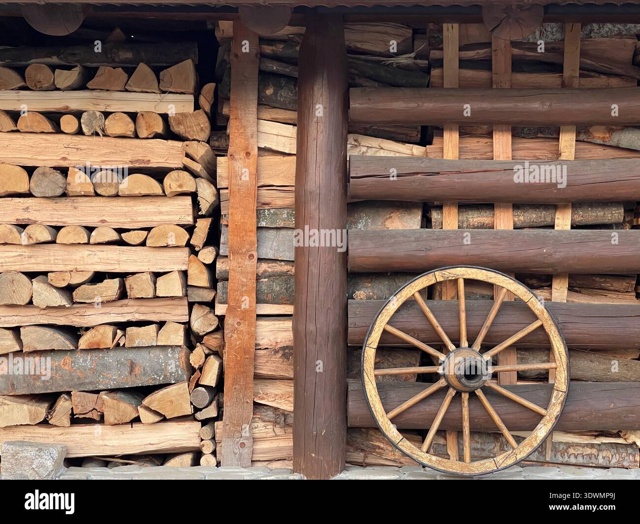 A decorative wooden wagon wheel leaning against a wall of neatly stacked firewood and logs. - Smartphone Captured Stock Image