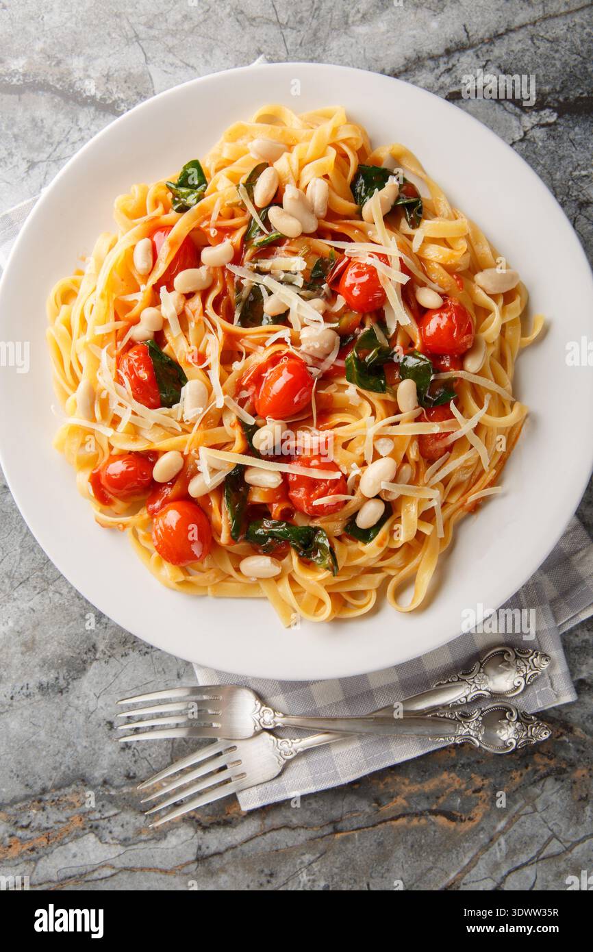 Italian tagliatelle pasta served on a white plate with roasted cherry tomatoes, white beans, spinach and grated parmesan closeup on the plate on the t Stock Photo