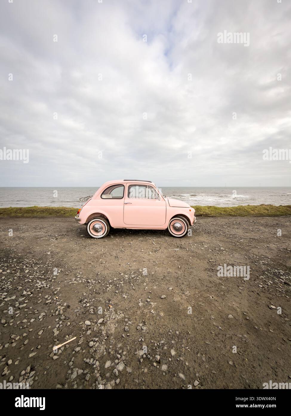 Vintage light pink Fiat 500 parked on a gravel road near the moving ocean waves under an overcast sky - Smartphone Captured Stock Image