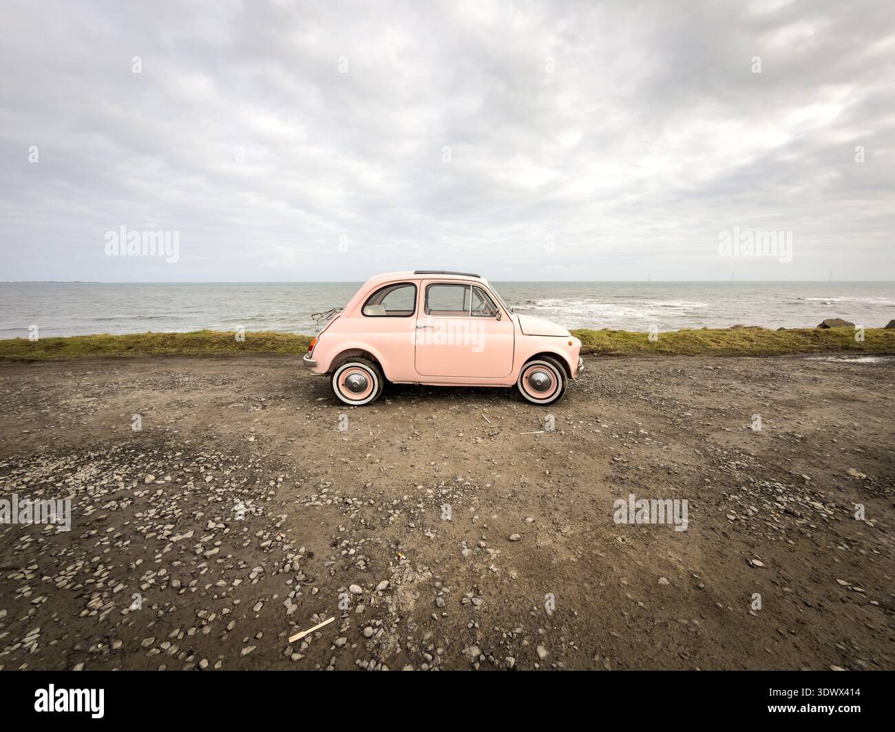 Vintage light pink Fiat 500 parked on a gravel road near the moving ocean waves under an overcast sky - Smartphone Captured Stock Image