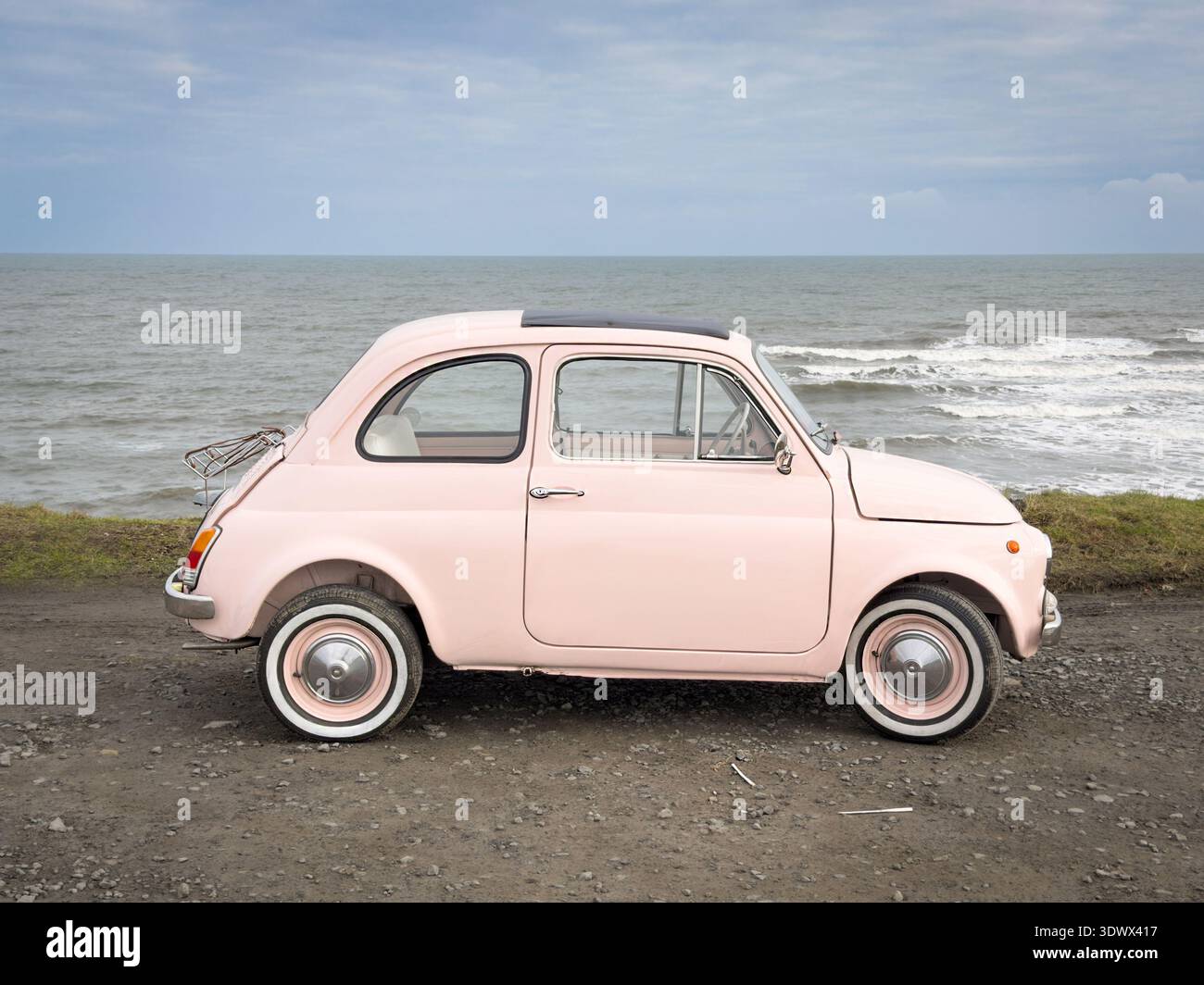 Vintage light pink Fiat 500 parked on a gravel road near the moving ocean waves under an overcast sky - Smartphone Captured Stock Image