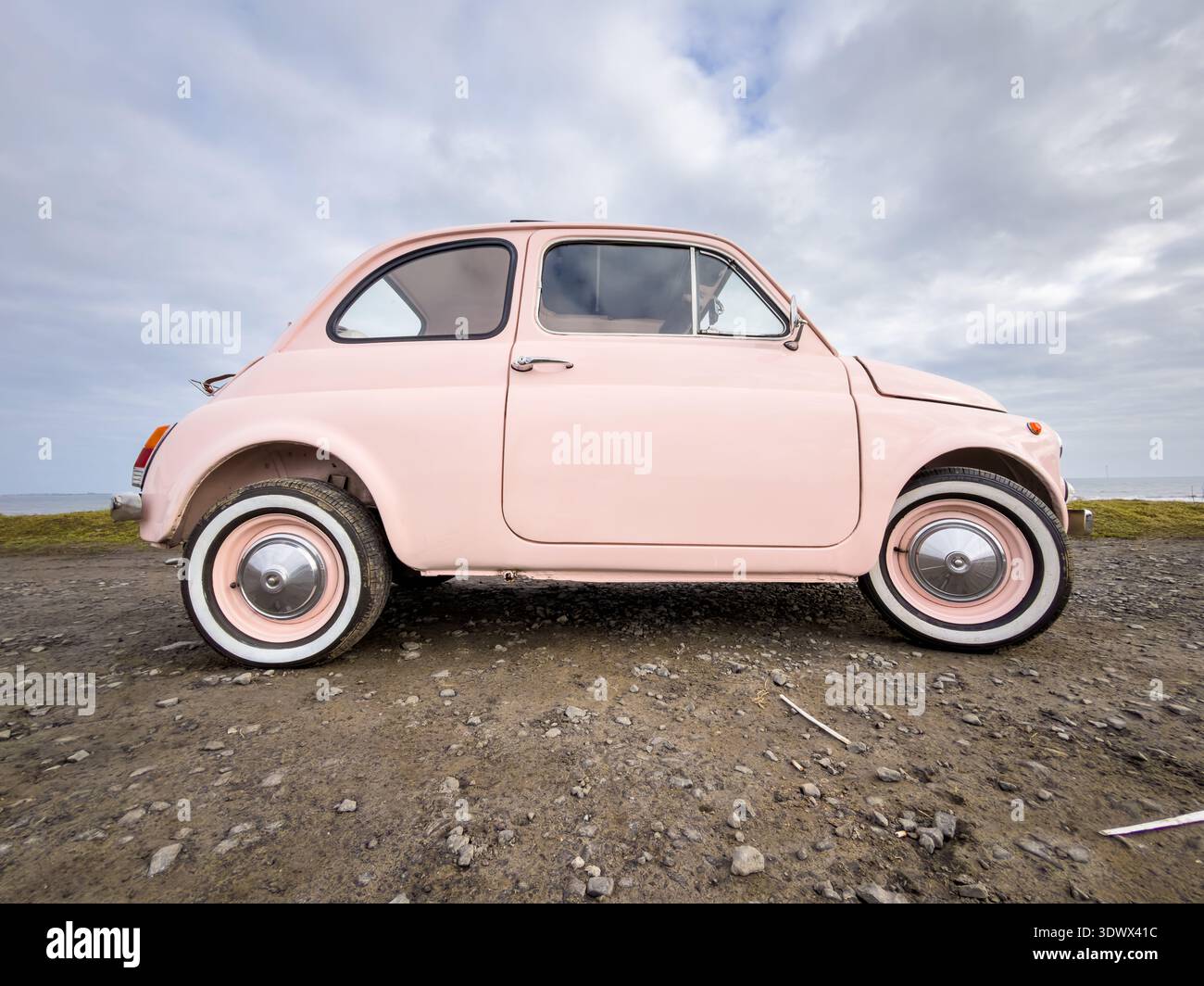 Side profile of a vintage light pink Fiat 500 car parked on a dirt road with an overcast horizon - Smartphone Captured Stock Image
