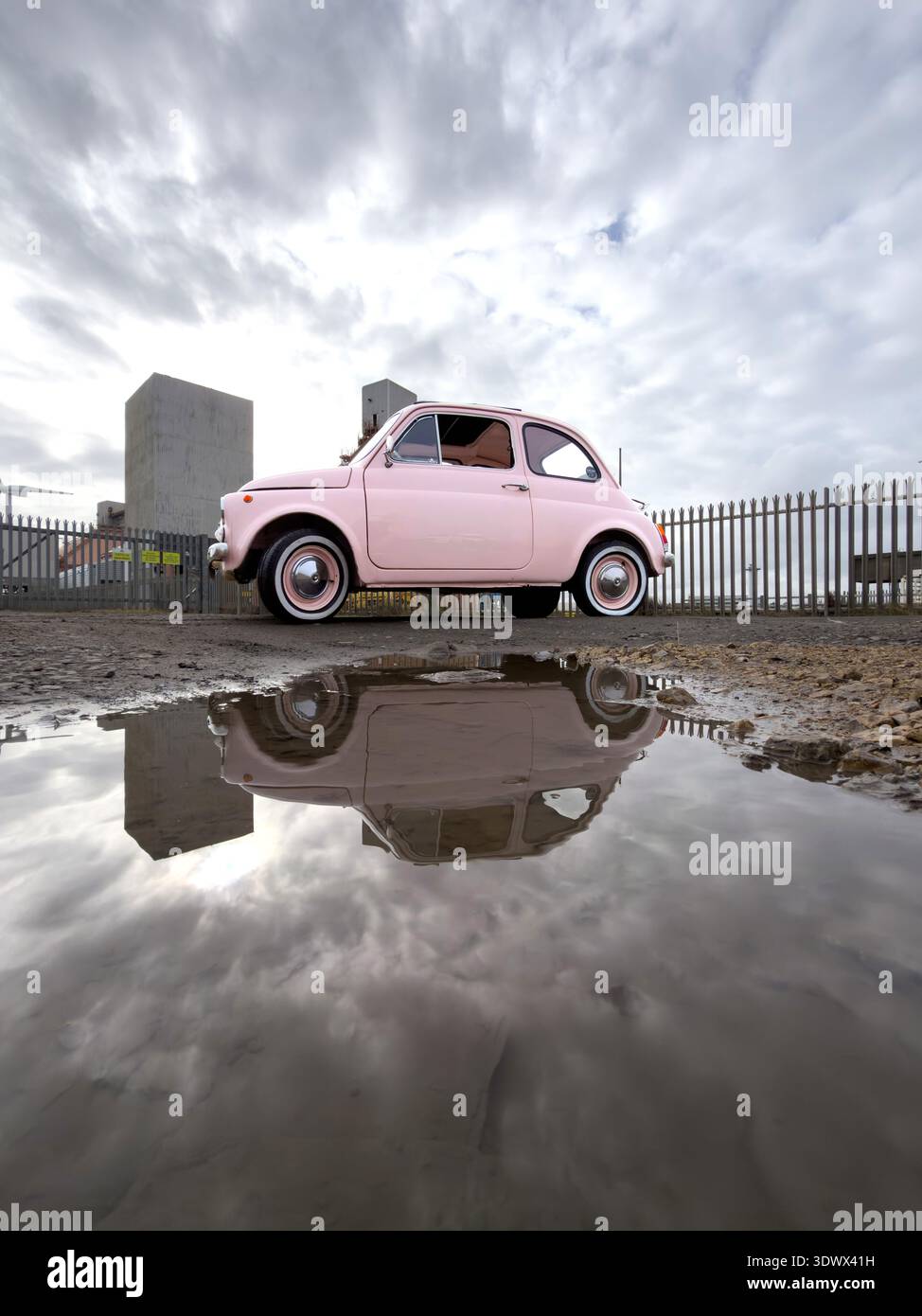 Pink classic Fiat 500 reflected in a large puddle under a dramatic overcast sky with industrial elements blyth northumberland uk - Smartphone Captured Stock Image