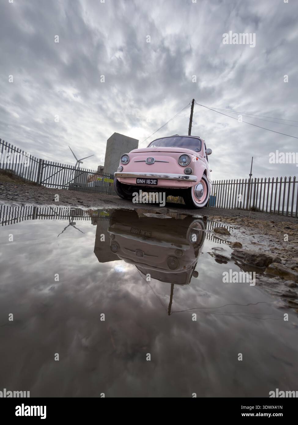 Pink classic Fiat 500 reflected in a large puddle under a dramatic overcast sky with industrial elements blyth northumberland uk - Smartphone Captured Stock Image