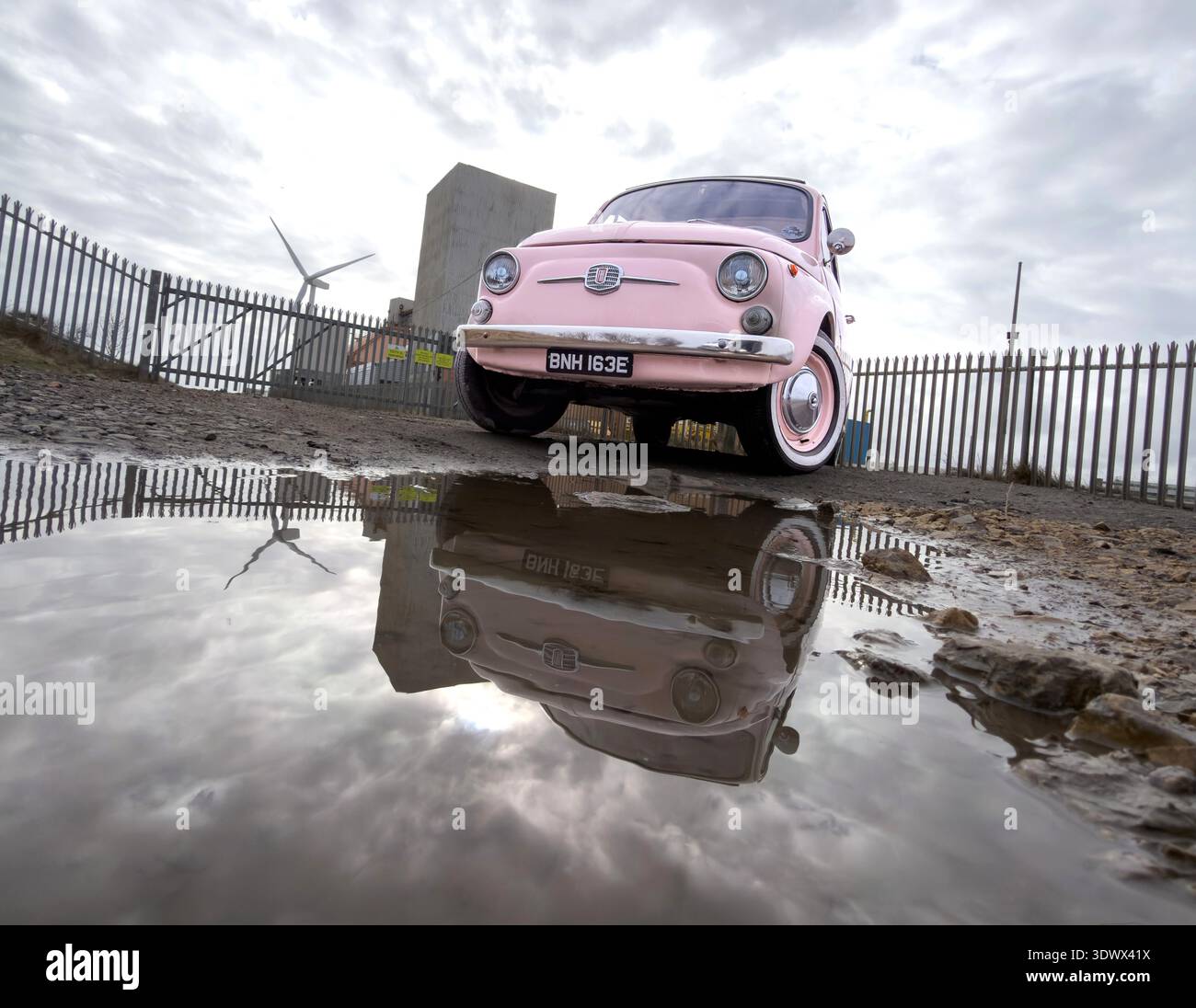 Pink classic Fiat 500 reflected in a large puddle under a dramatic overcast sky with industrial elements blyth northumberland uk - Smartphone Captured Stock Image