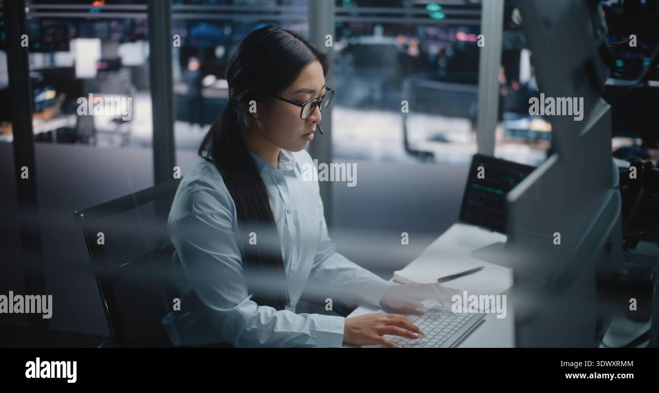 Asian Female Financial Specialist Sits at Innovative Workspace, Eyes Scanning Multiple Displays Filled With Trading Indices. Young Lady Actively Uses Keyboard to Input Data and Execute Market Orders. Stock Photo