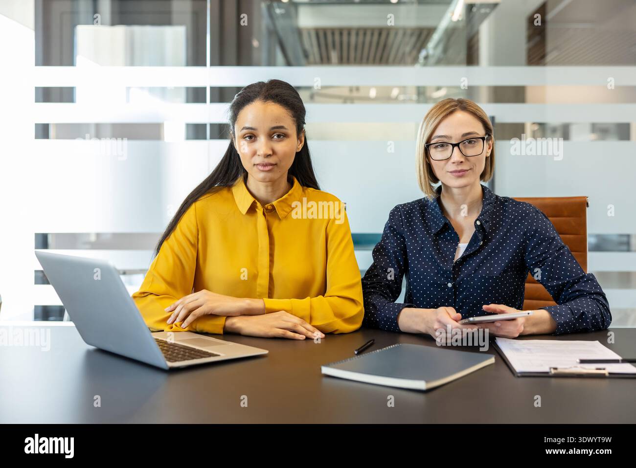 Two diverse businesswomen are sitting at a conference table with a laptop, tablet, and documents, actively engaging in professional collaboration within a modern glass-partitioned office environment Stock Photo
