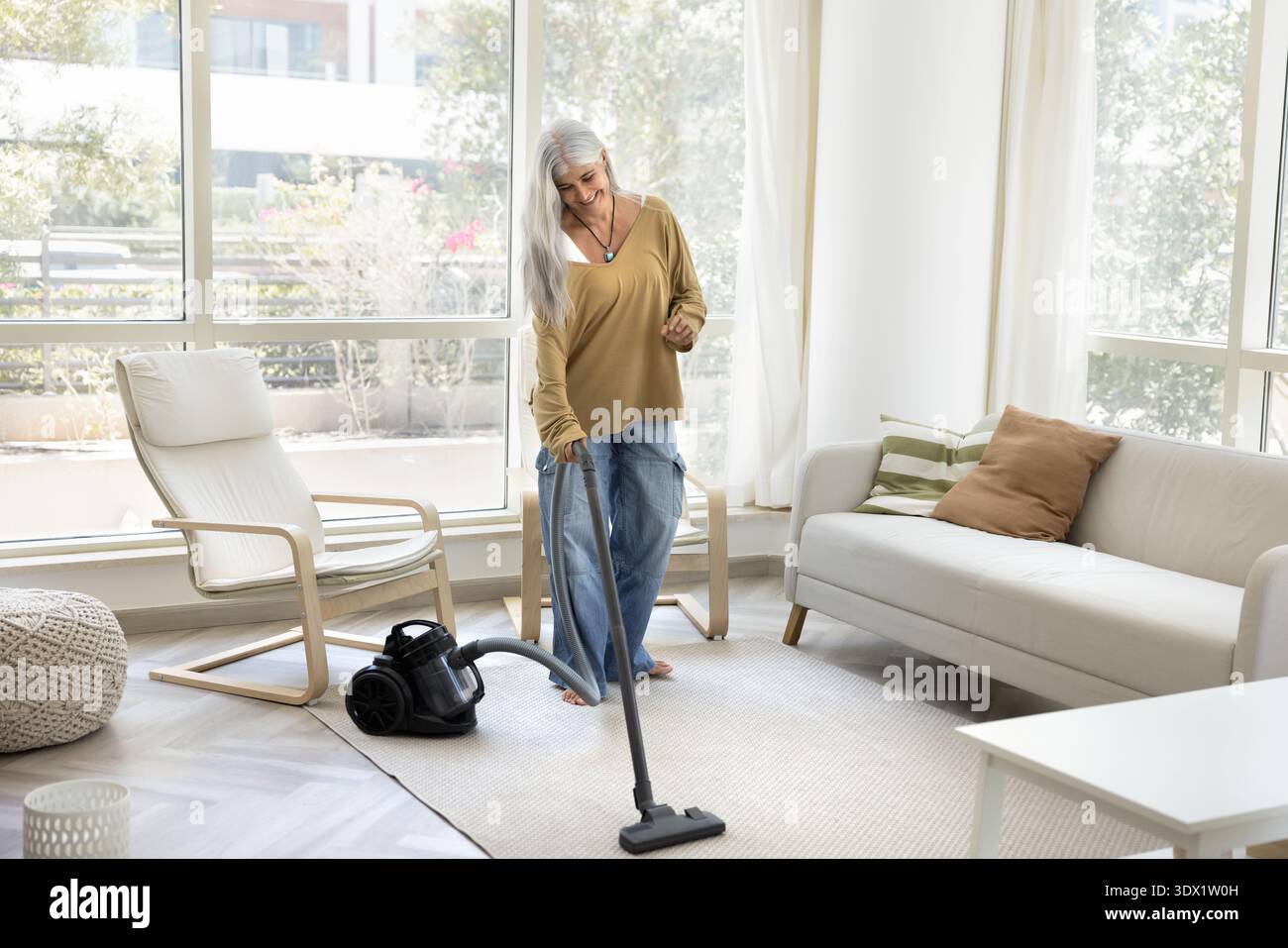 Smiling mature woman with long silver hair vacuuming living room Stock Photo
