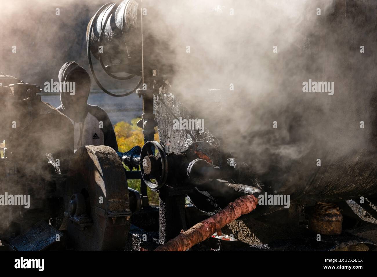 A hooded man operates a soot-covered bitumen boiler amidst thick smoke during road construction work in the village of Sagnam. Stock Photo