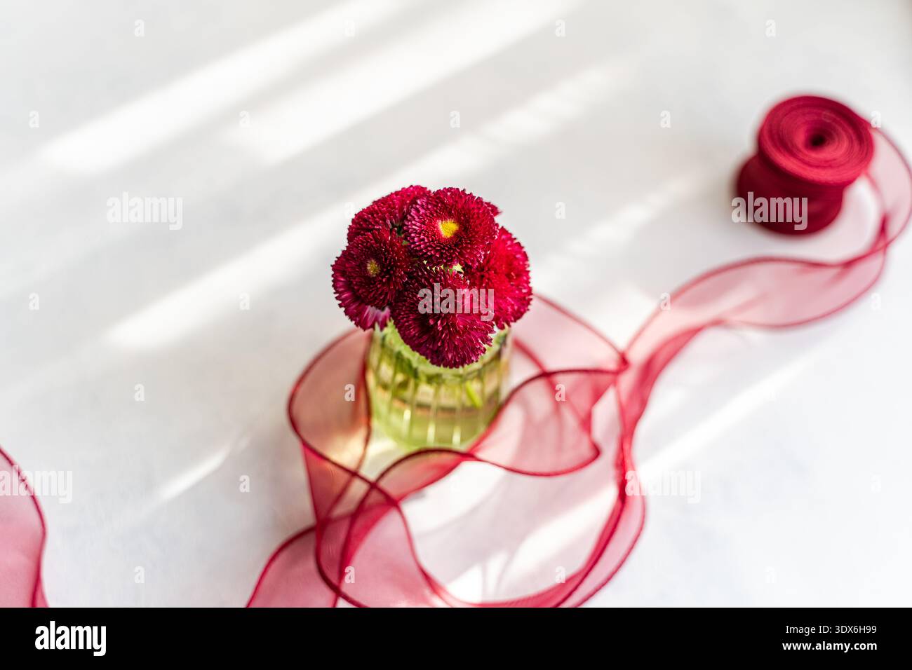 A bright spring scene featuring vibrant red flowers in a vase, accompanied by a delicate red ribbon elegantly spread across a white table, creating a Stock Photo