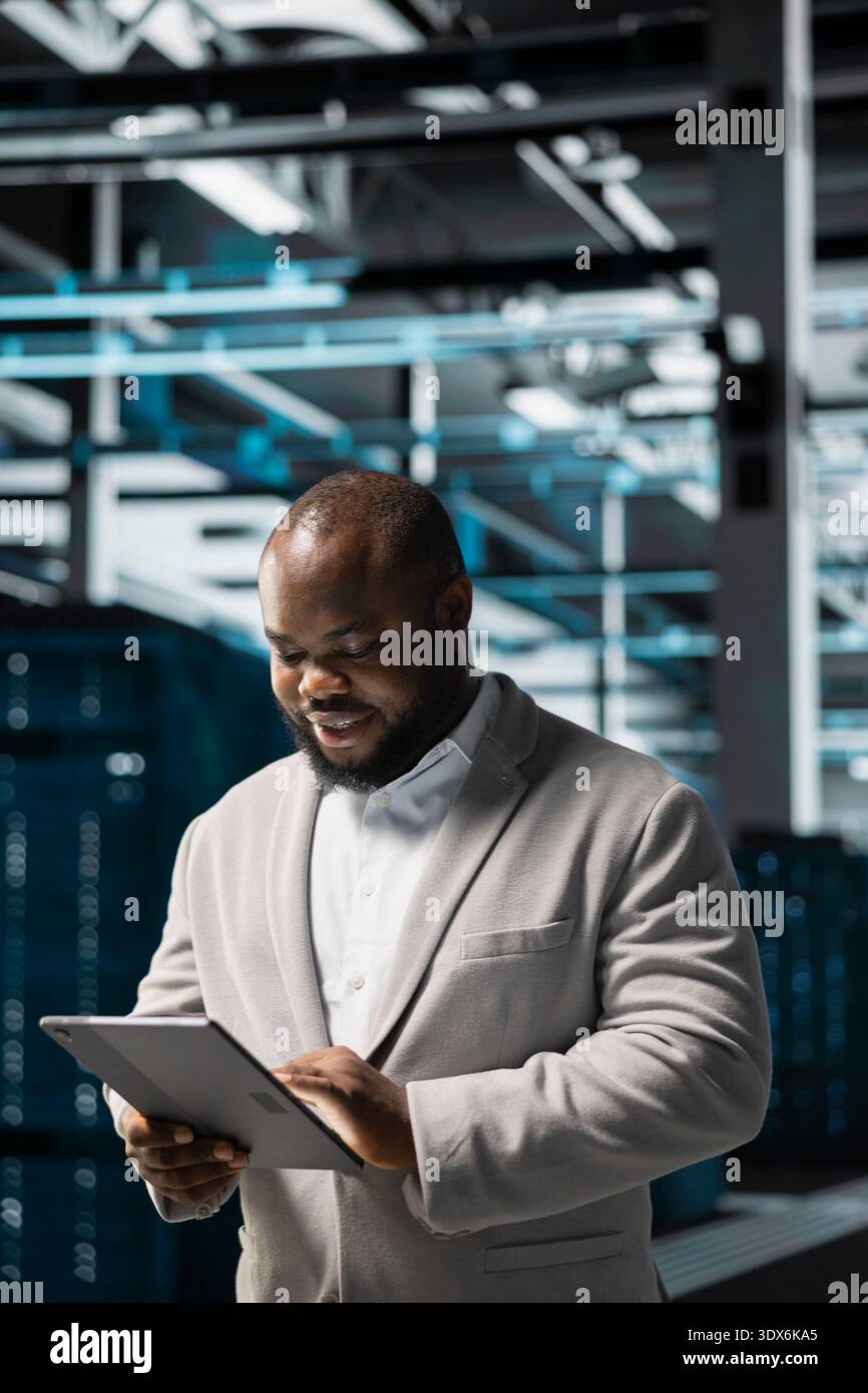 Cheerful technician in data center workspace running system diagnostic tests on tablet to fix problems. Joyous employee looking for network issues affecting rigs performance using device Stock Photo