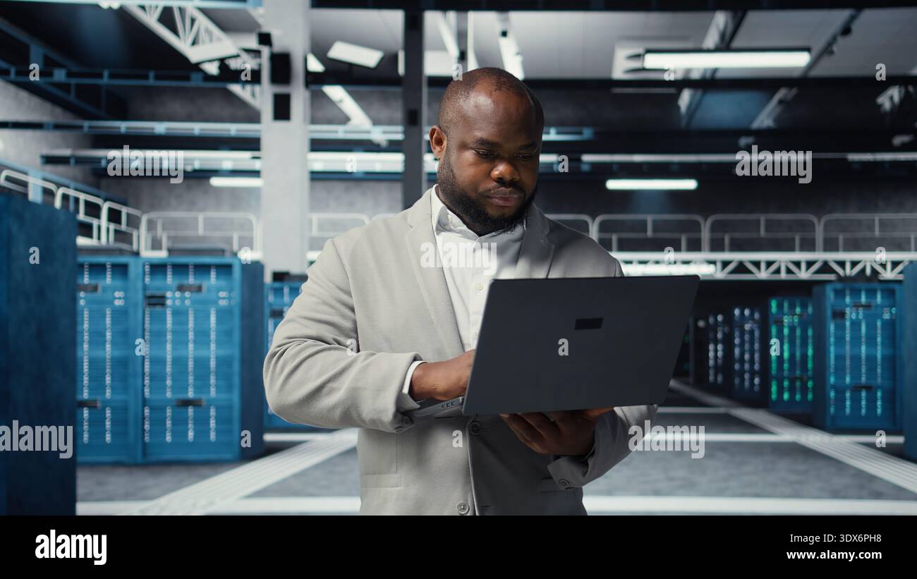 Technician walking in data center, using laptop to check hardware for system stability. Worker overseeing server farm, using notebook to configure infrastructure, optimizing performance, camera A Stock Photo