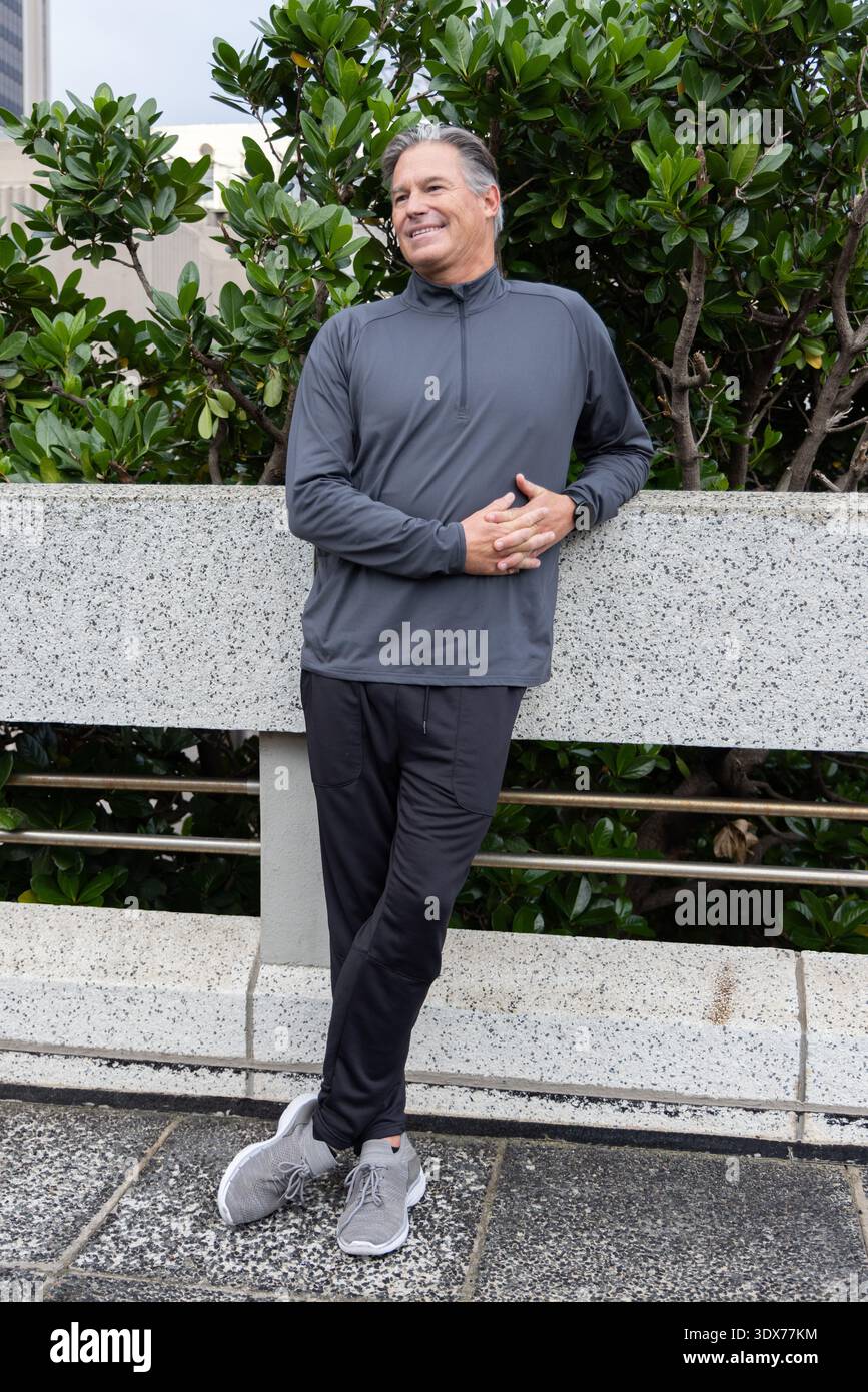 Middle-aged man standing and leaning against speckled planter wall on urban plaza, wearing pullover Stock Photo