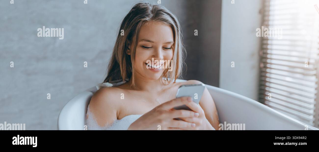 Woman Using Smartphone While Relaxing in Bathtub With Natural Light in Background Stock Photo