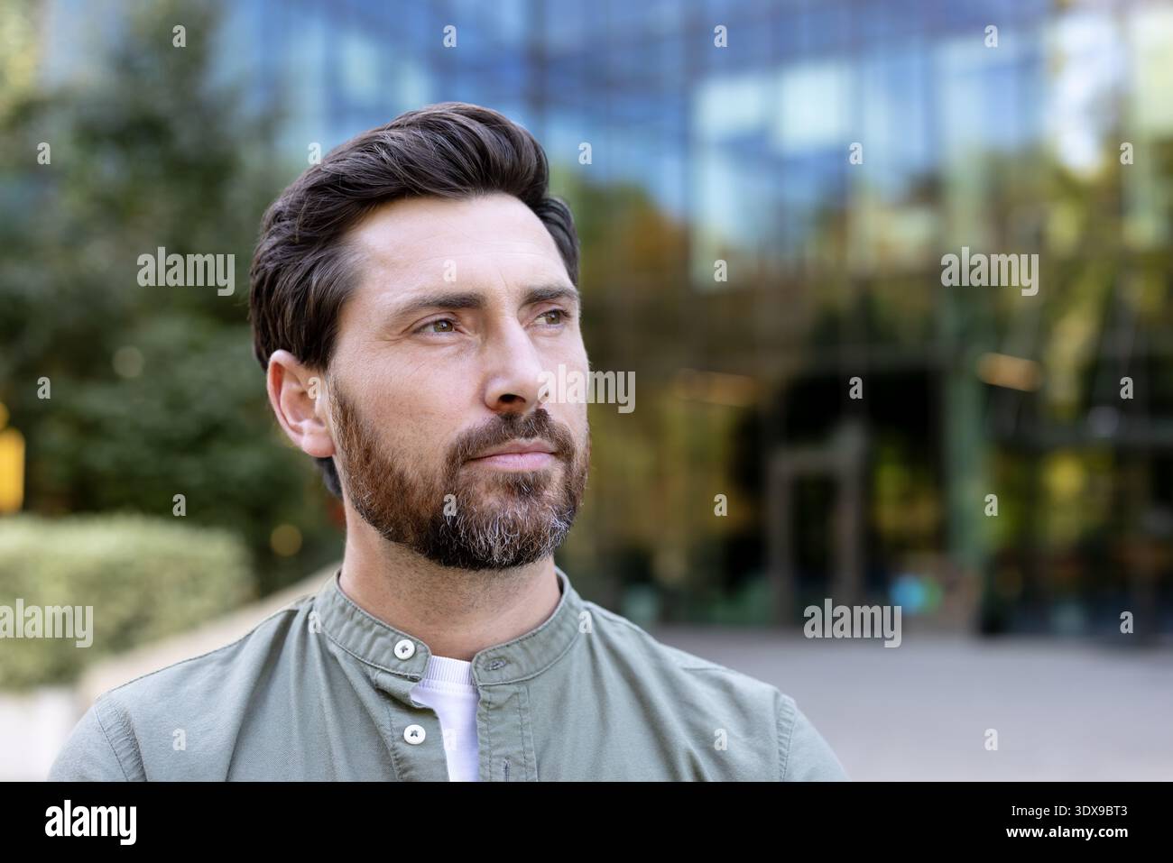 Bearded man with a serious, pensive expression looks away while standing outdoors against a modern office building, conveying confidence, determination and urban ambition Stock Photo