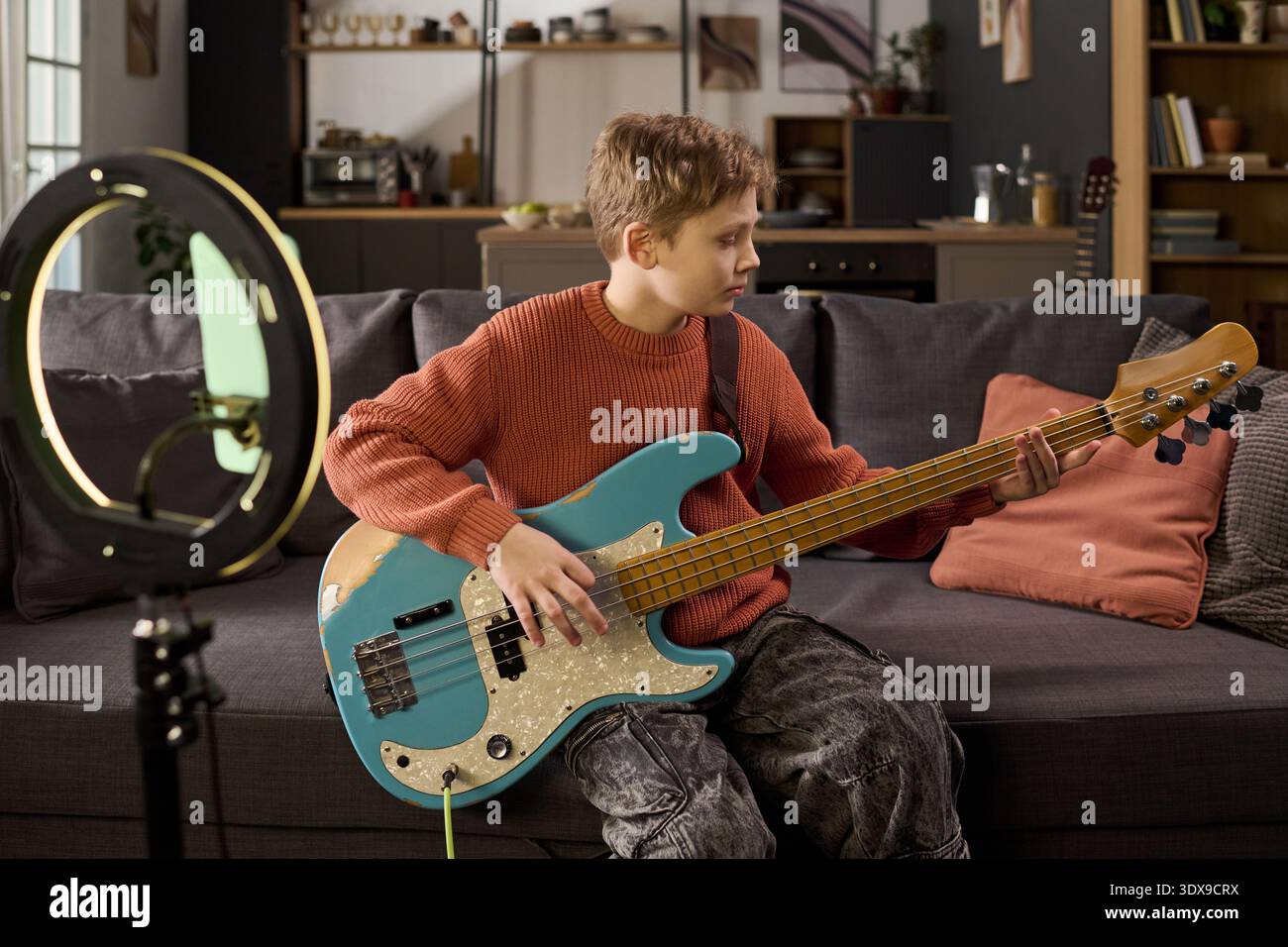 Caucasian boy playing electric bass guitar sitting on sofa, focusing on instrument, ring light positioned in foreground, home interior visible in background, casual setting Stock Photo
