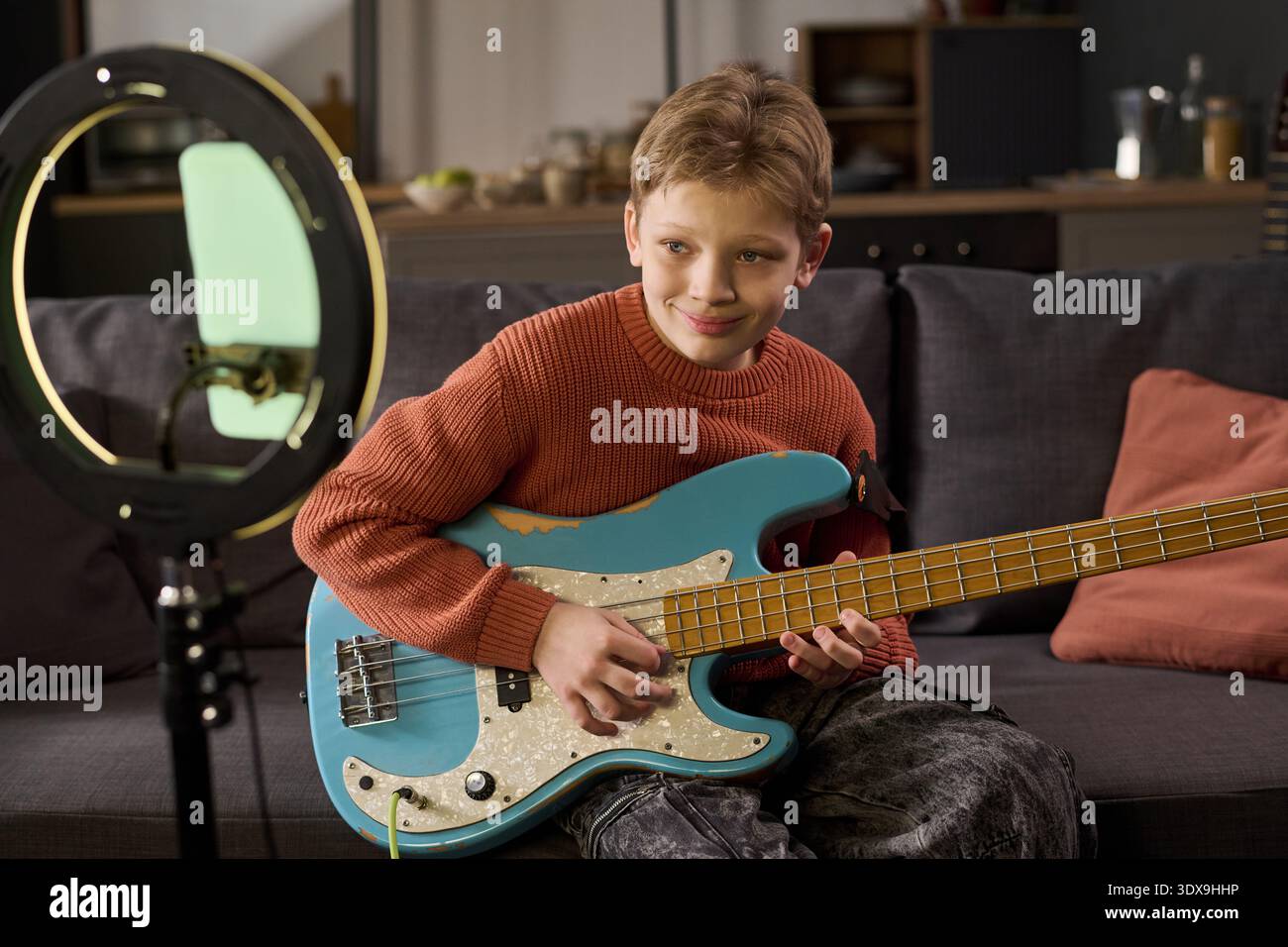 Caucasian boy playing electric bass guitar while sitting on sofa, smiling and looking slightly away from camera, recording music video using smartphone mounted on ring light Stock Photo